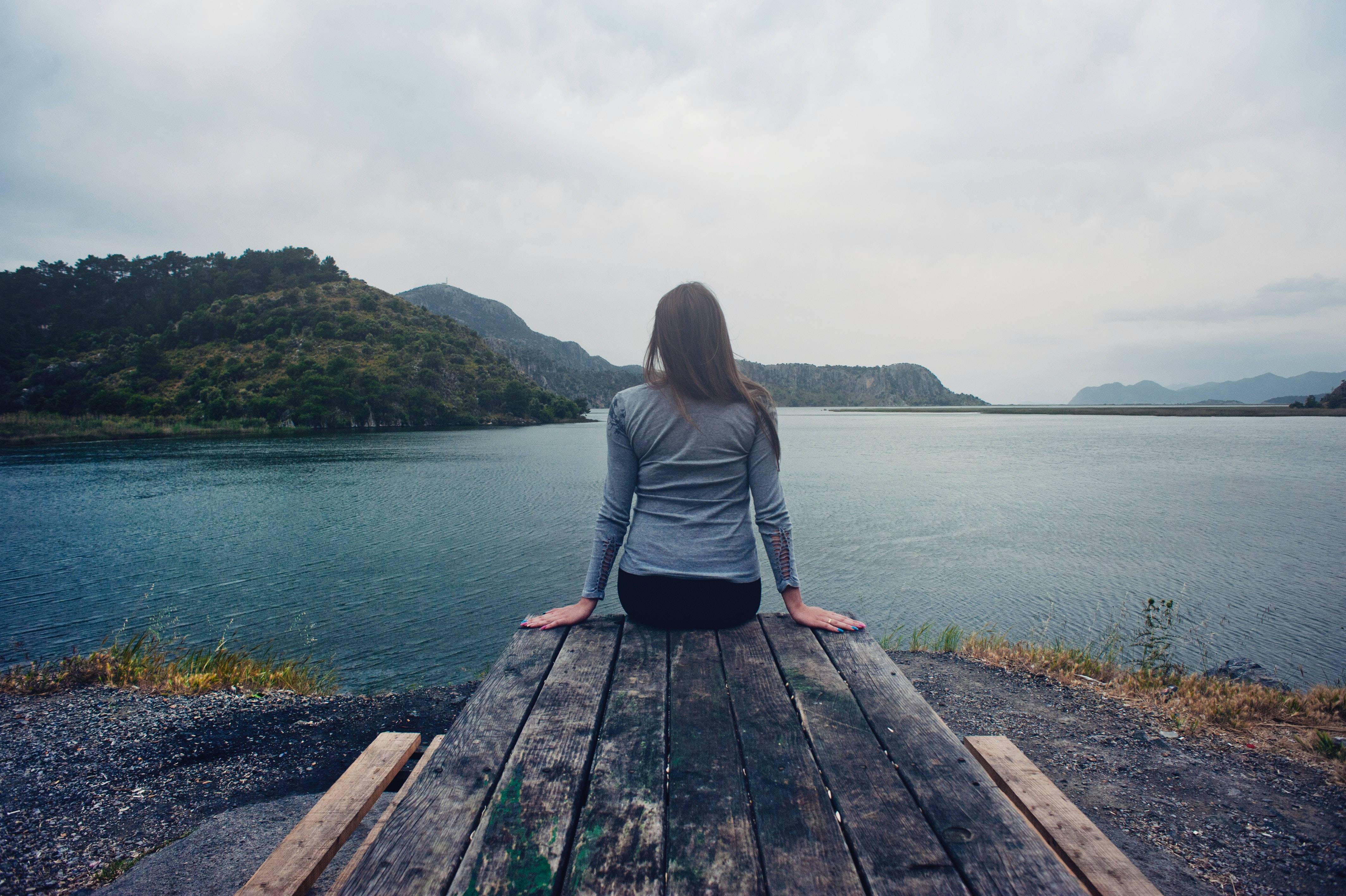 Sad Girl 桌布 壁紙 — 一個人獨自坐在一個經風化的木碼頭上，俯瞰著一個被森林覆蓋的山丘環繞的平靜藍色湖泊，天空多雲。
