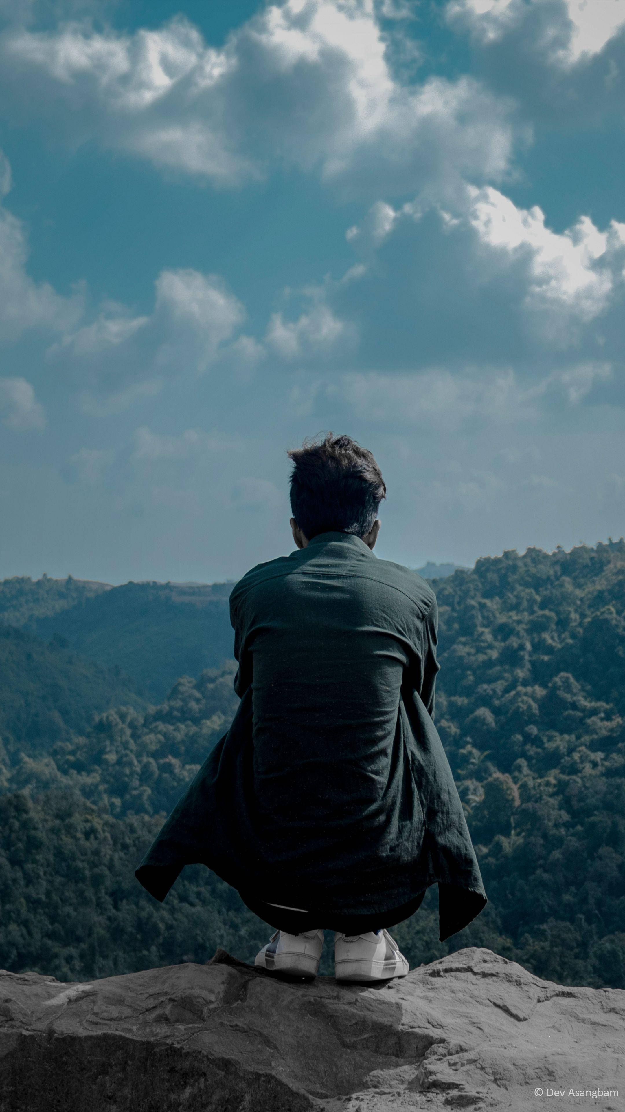 Alone Boy 桌布 壁紙 — 一個人獨自坐在岩石懸崖邊緣，俯瞰著廣闊的森林山谷，天空烏雲密佈，氣氛戲劇化。