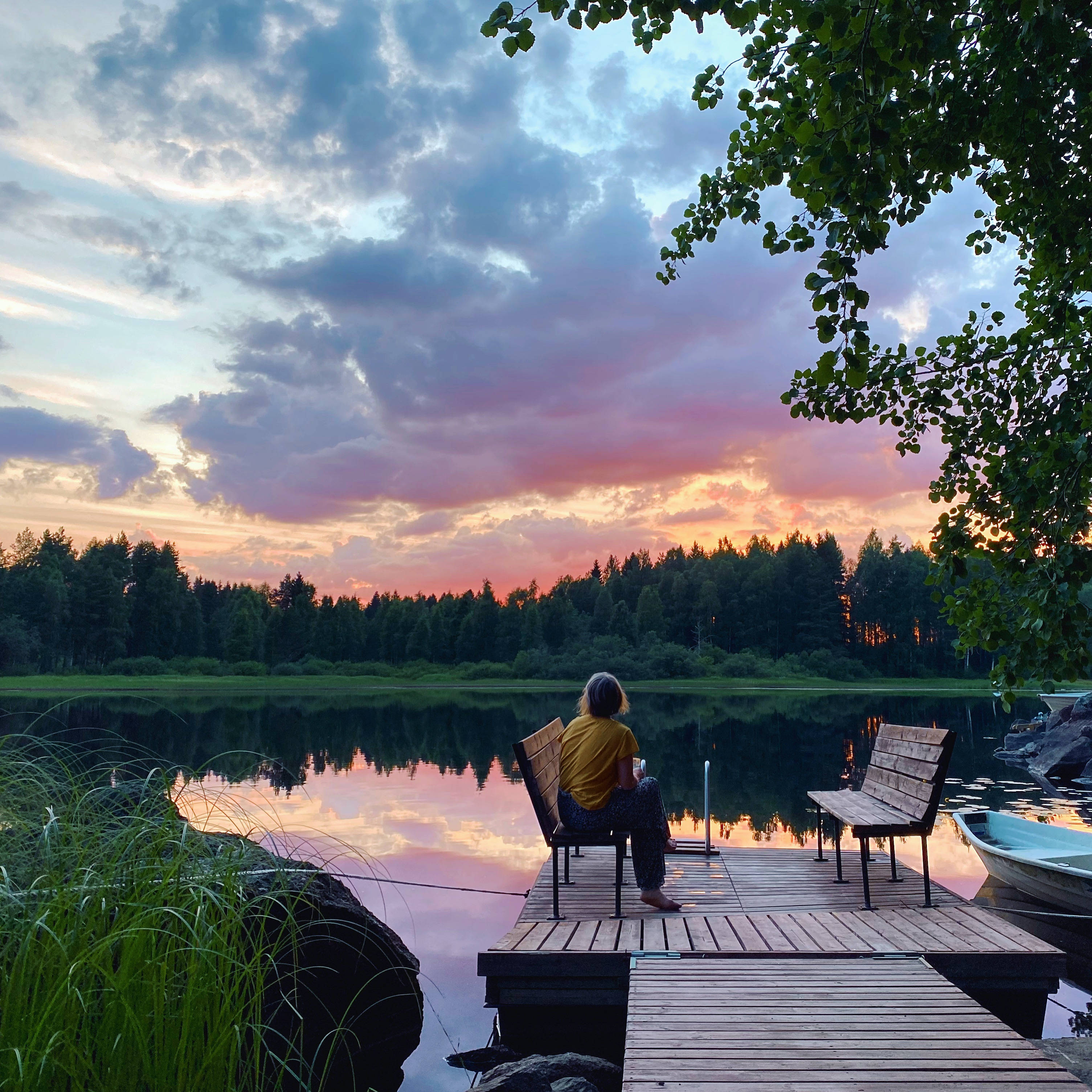Finland 桌布 壁紙 — 一個人獨自坐在日落時分的木碼頭上，觀賞樹木環繞的平靜湖水中倒映出鮮豔的粉紅色和橙色雲彩。