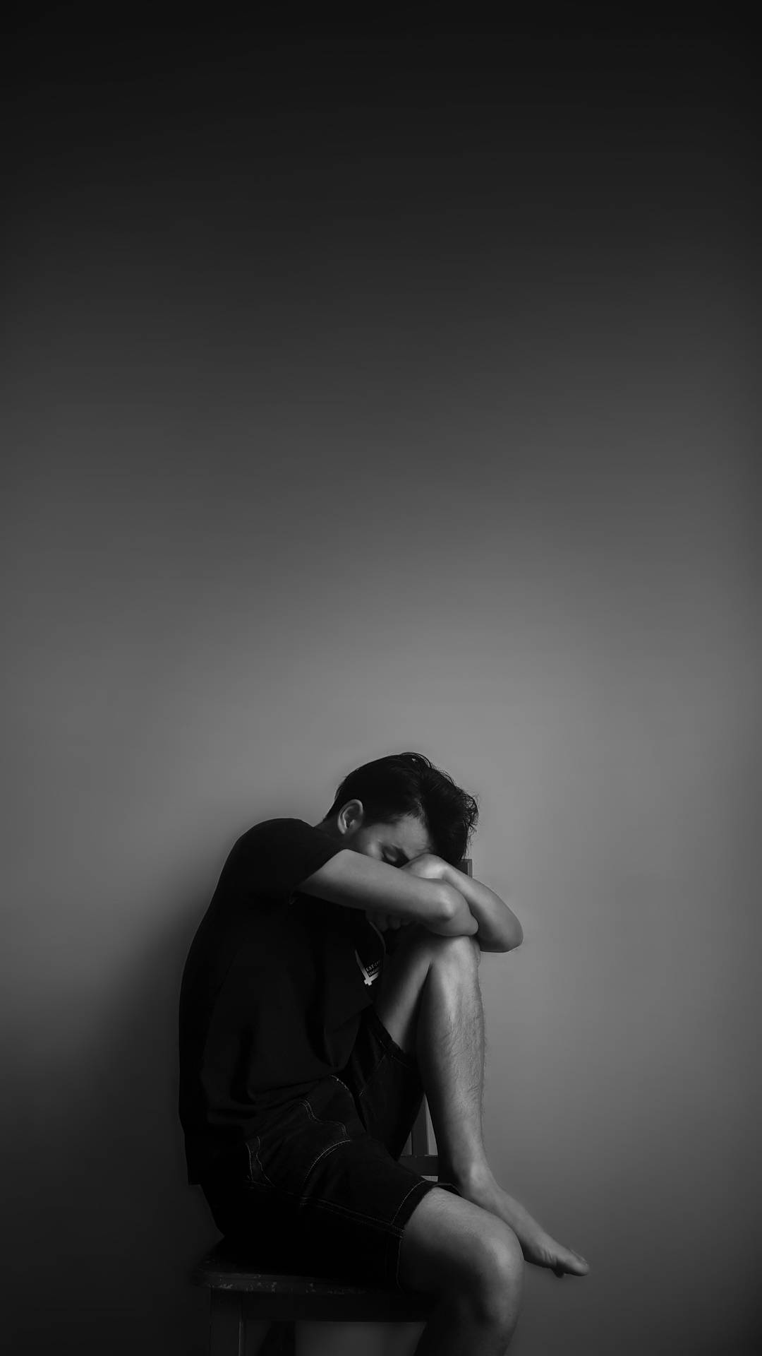 一個人獨自坐在凳子上，低著頭，在灰階房間中。 — 憂鬱男孩 壁紙