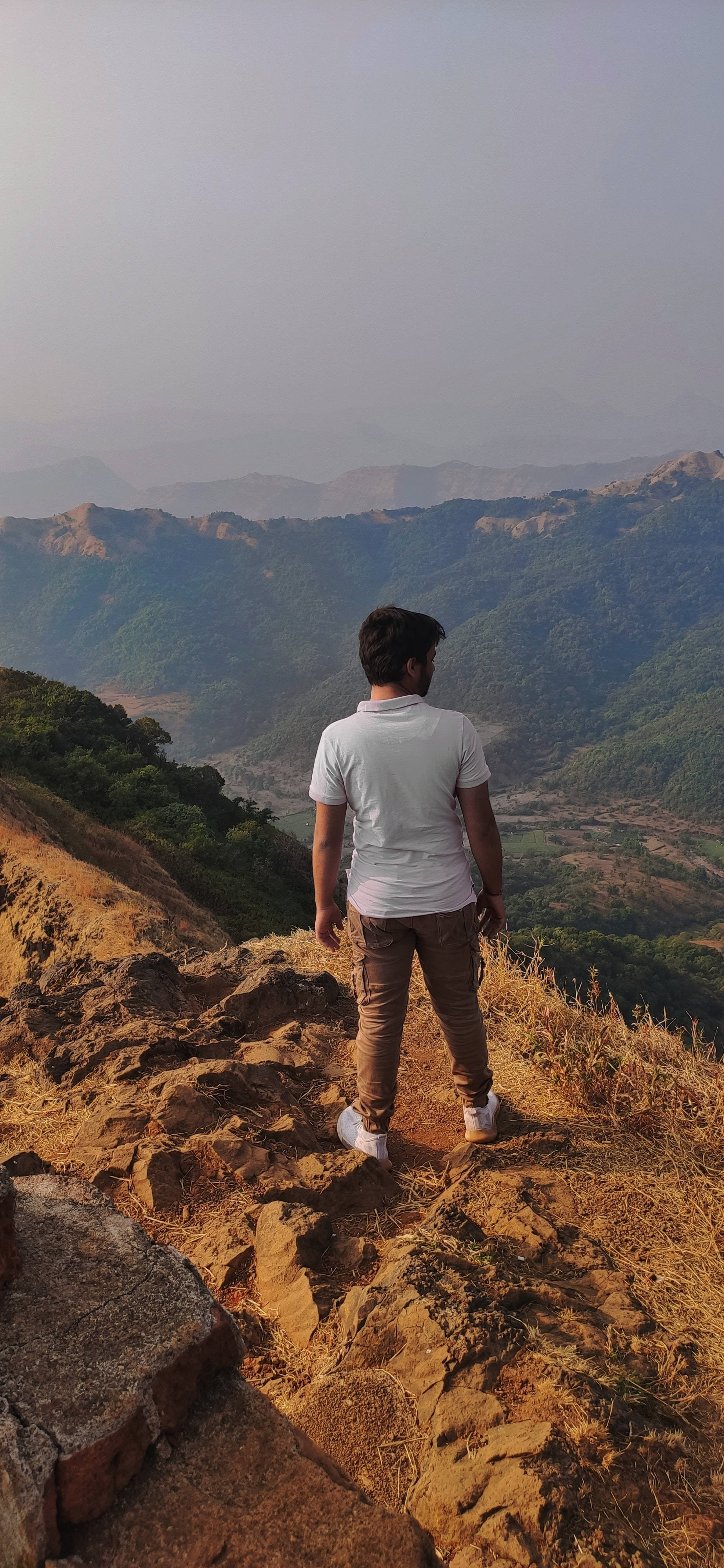Scenery 桌布 壁紙 — 一個人獨自站在岩石山脊上，俯瞰著廣闊的霧氣山谷，層層疊疊的綠色山丘在柔和天空下延伸。