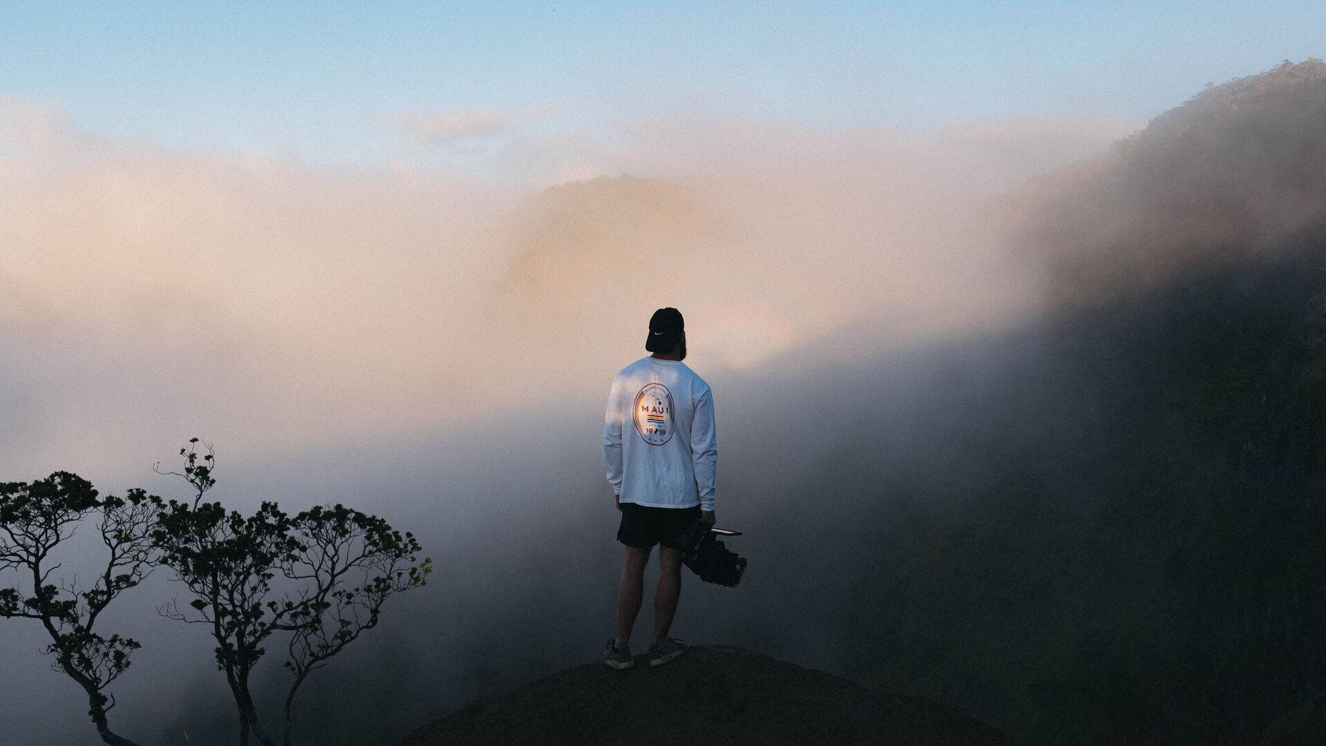 Travel Desktop 桌布 壁紙 — 一個人獨自站在黎明時分霧氣籠罩的山頂上，俯瞰著雲海，附近有樹木的剪影。