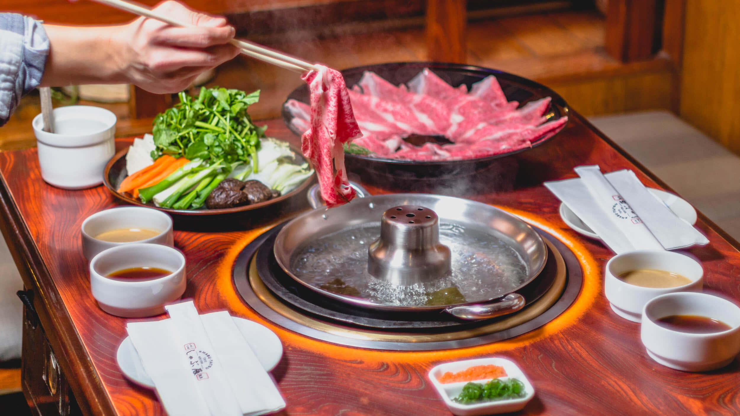 meat 桌布 壁紙 — 一個人用筷子將薄切牛肉提起，放在一個冒著熱氣的鍋上，周圍是新鮮蔬菜和蘸醬，放在木桌上。