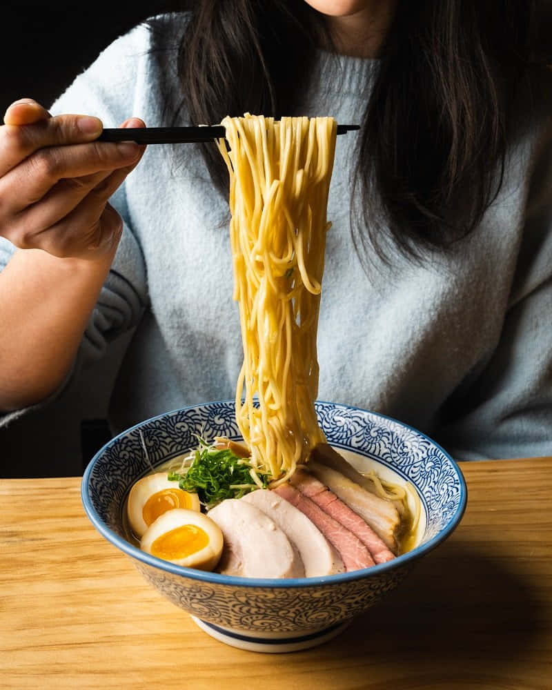 Noodles 桌布 壁紙 — 一個人用筷子從一個有圖案的碗中提起冒煙的拉麵，碗裡有湯、豬肉片、溏心蛋和蔬菜。