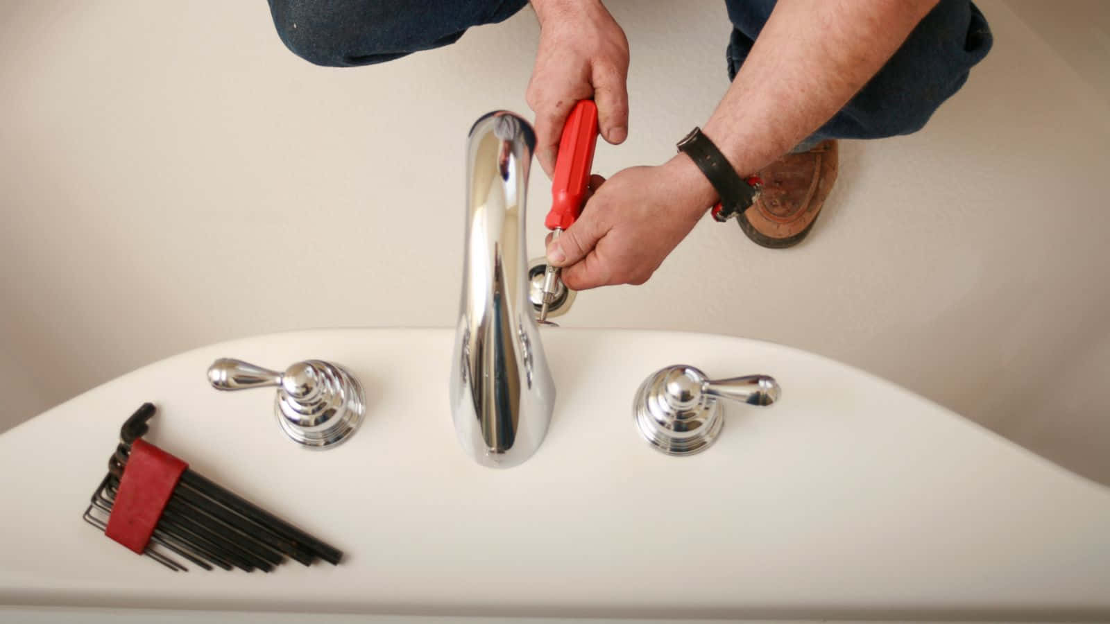 Plumbing 桌布 壁紙 — 一個人用紅色鉗子收緊白色洗手台上的鉻水龍頭，附近有工具。