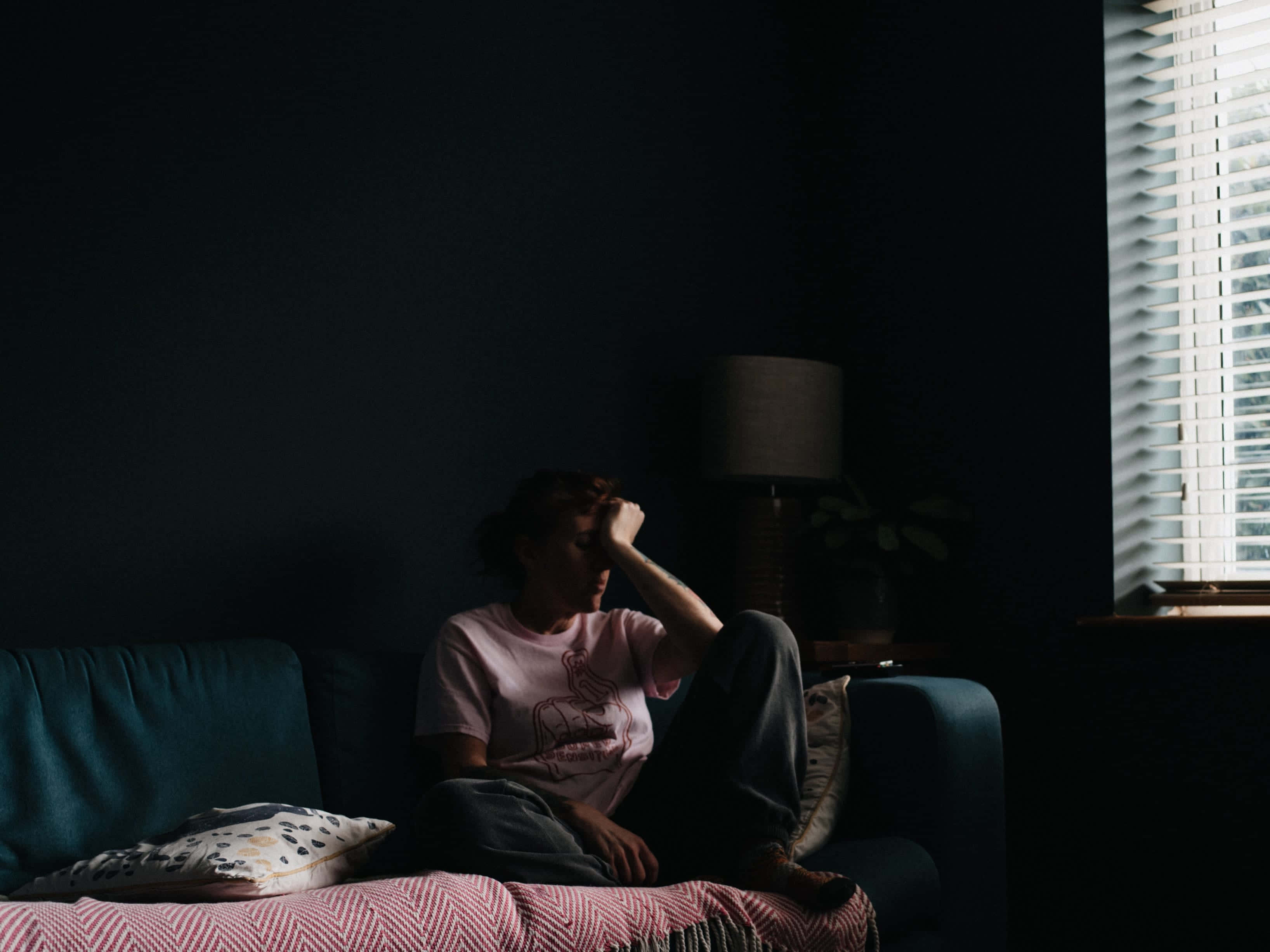 Lonely 桌布 壁紙 — 一個人癱坐在昏暗房間的沙發上，手按額頭，沐浴在帶百葉窗的窗戶透進的柔和光線中。