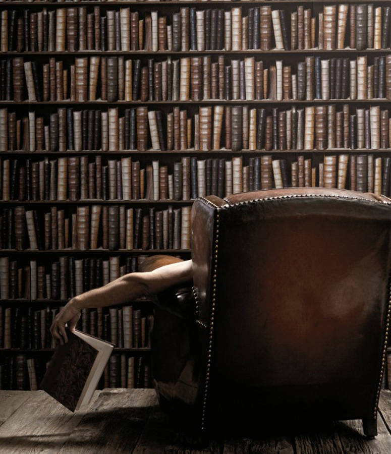 chair 桌布 壁紙 — 一個人癱軟在皮革扶手椅上，拿著一本書，背景是高聳的書架，放滿了復古書籍。