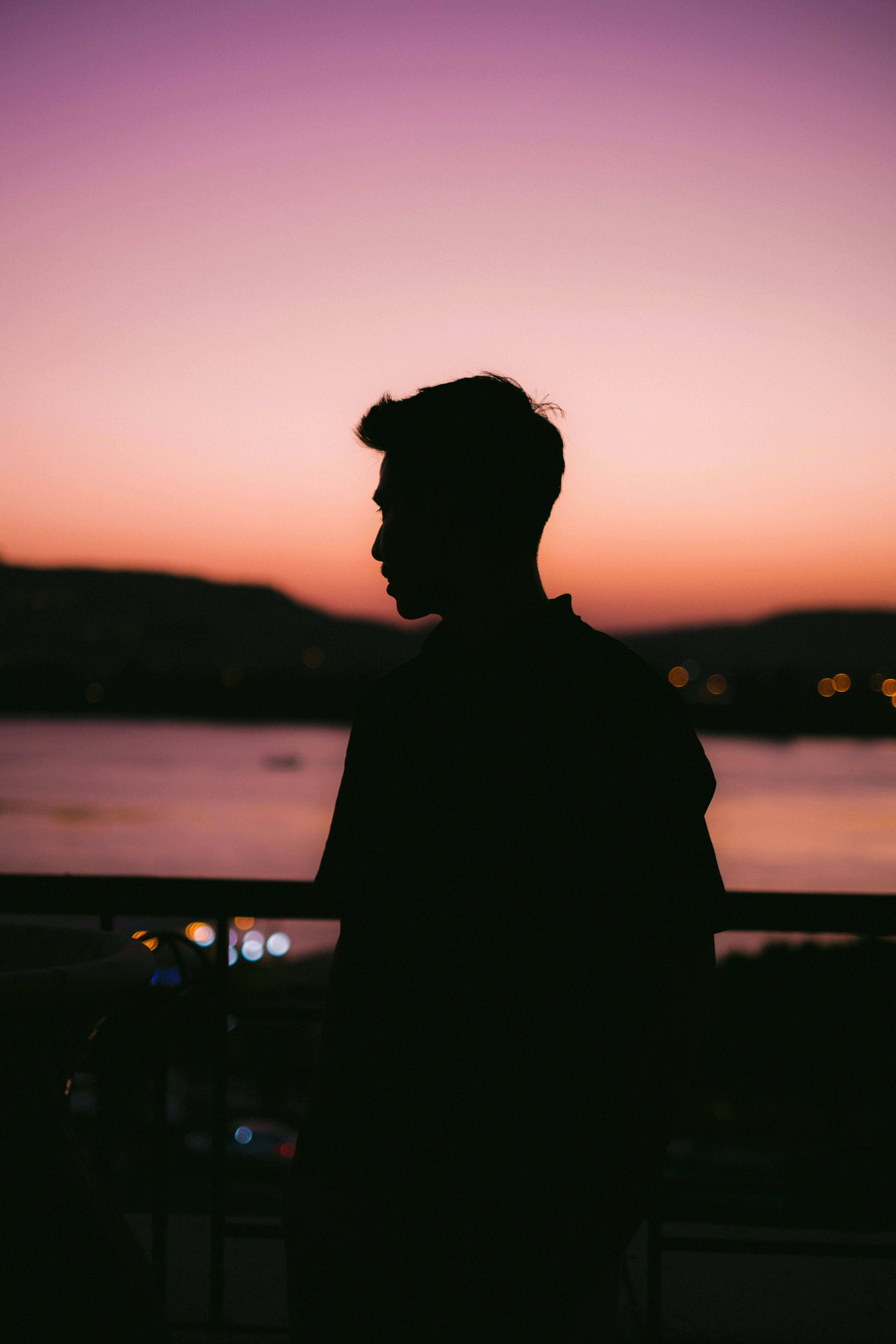 Man Aesthetic 桌布 壁紙 — 一個人的剪影，凝視著水面上充滿活力的紫色和橙色日落，遠方有城市燈光。