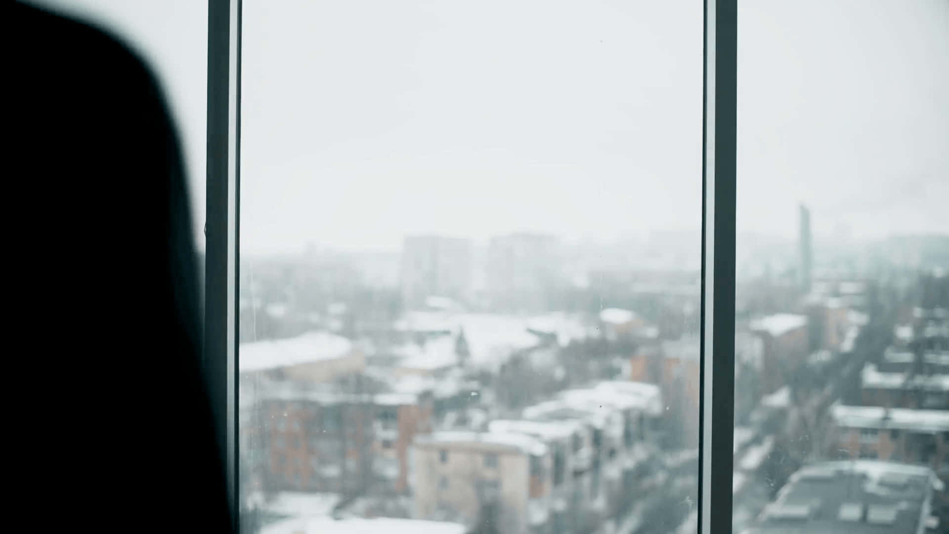 window 桌布 壁紙 — 一個人的剪影，在陰天時透過窗戶凝視著白雪覆蓋的城市景觀。