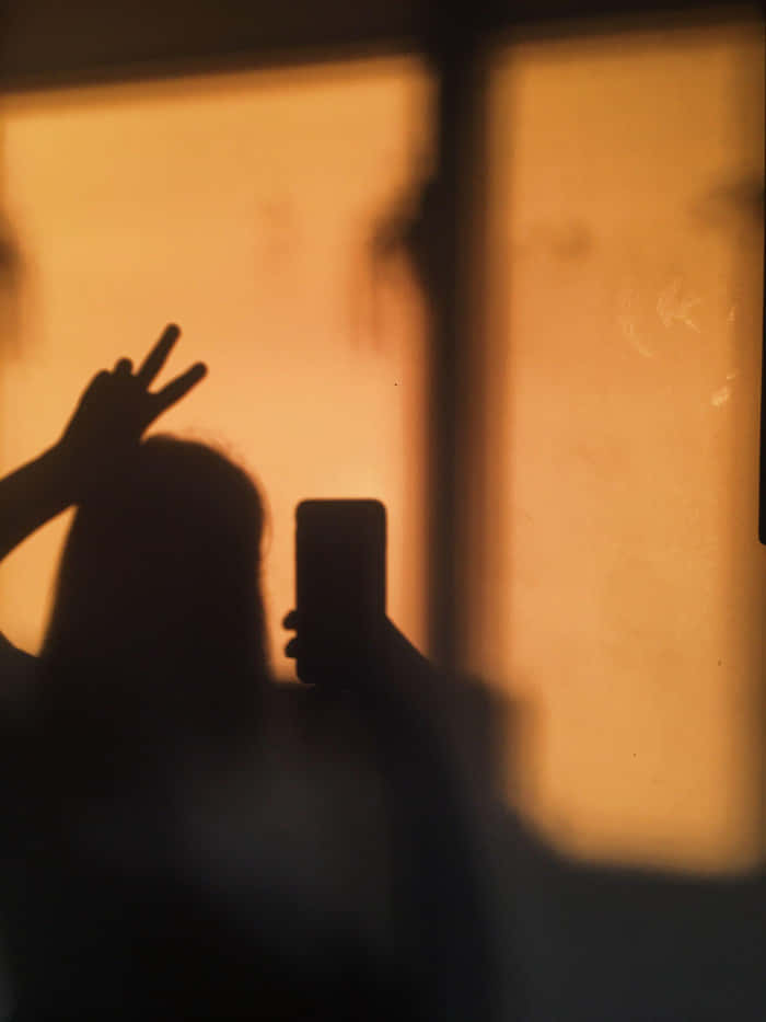 Girl Shadow 桌布 壁紙 — 一個人的剪影，手持電話並在溫暖發光的窗戶前做出和平手勢。
