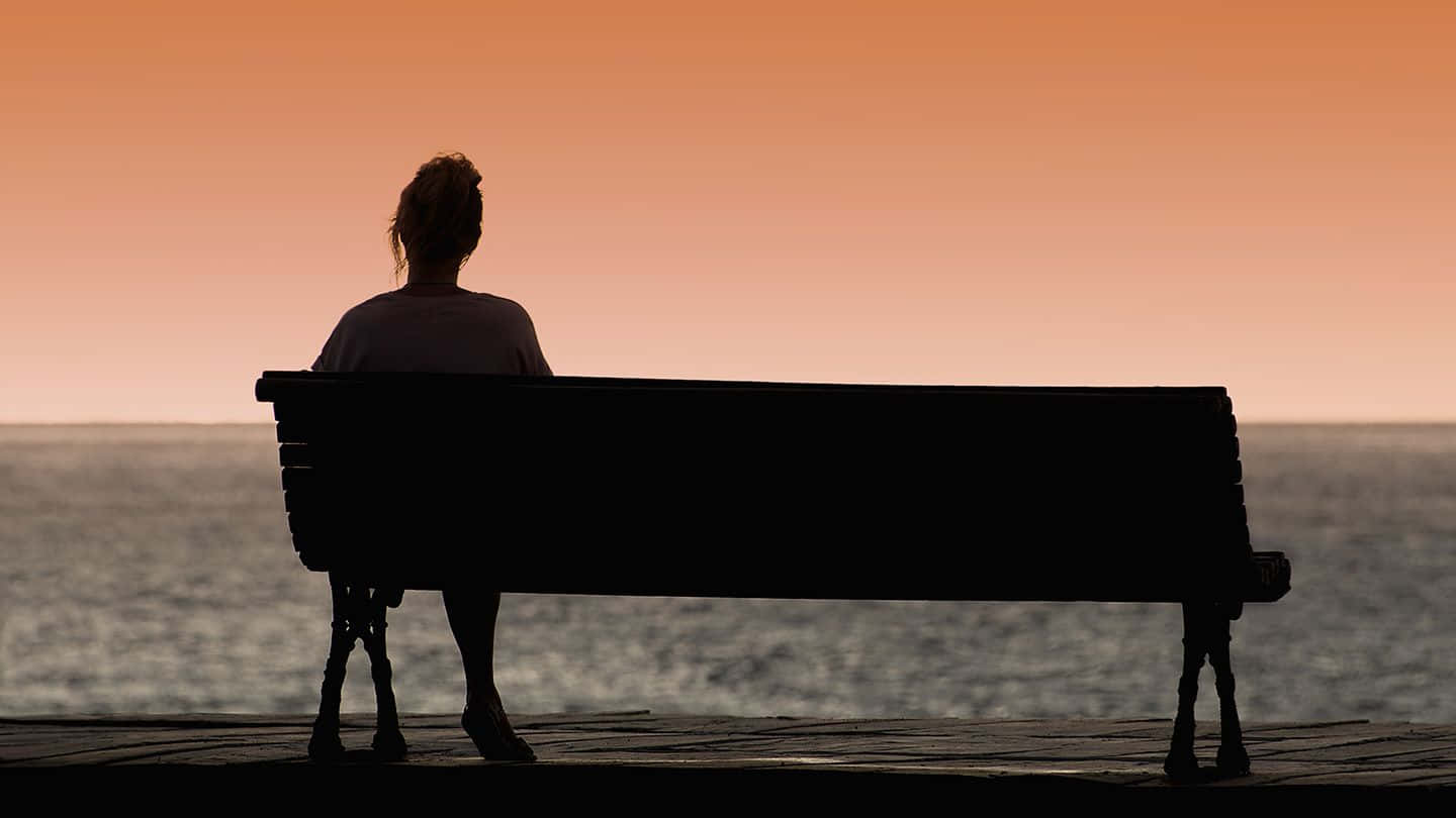 Lonely 桌布 壁紙 — 一個人的剪影，獨自坐在長椅上，面向平靜的海洋，夕陽西下，溫暖的橙色天空。