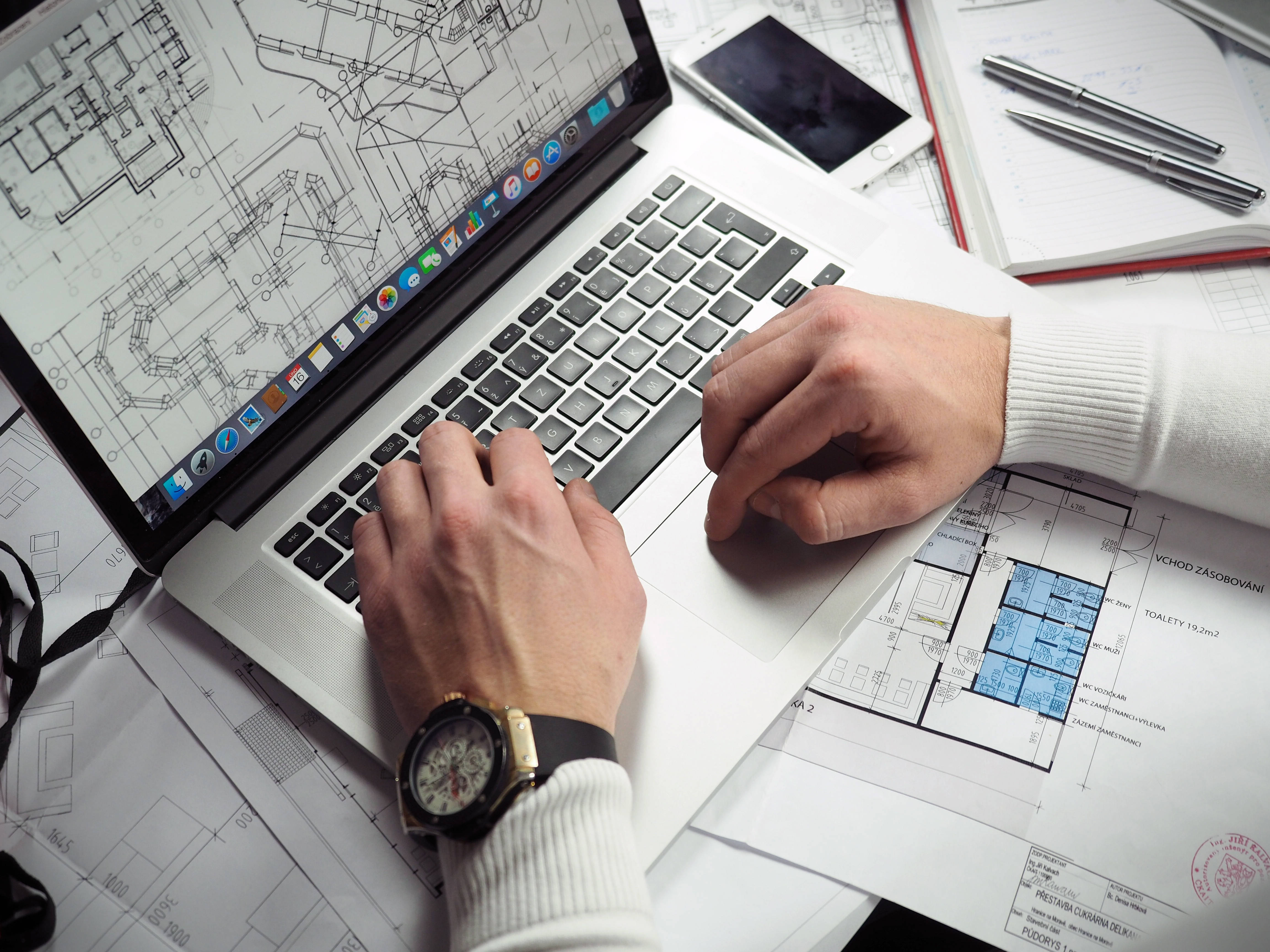 Working 桌布 壁紙 — 一個人的手在專注的工作空間中，在建築藍圖和設計工具環繞下於筆記型電腦上打字。