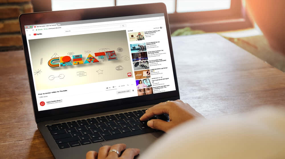 YouTube 桌布 壁紙 — 一個人的手在木桌上打字，筆記型電腦顯示一個關於用彩色3D字母創作內容的YouTube影片。