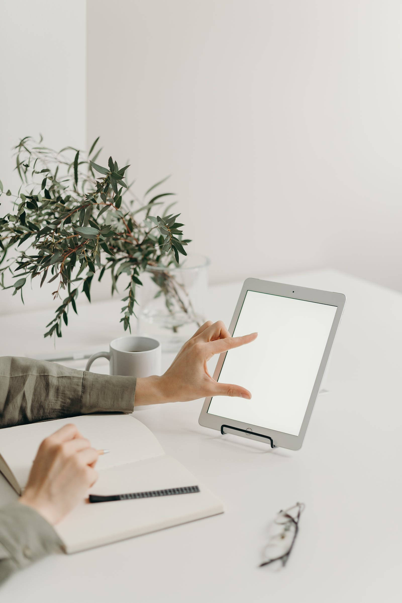 Apple iPad Pro 桌布 壁紙 — 一個人的手在簡約白色桌子上，旁邊放著綠色植物花瓶和馬克杯，一邊寫筆記本一邊操作平板電腦。