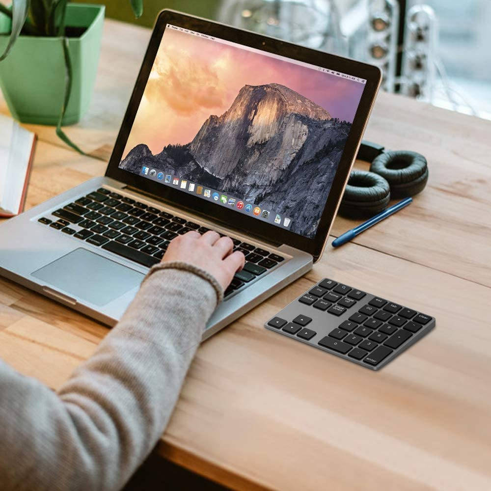 MacBook 動態圖片 桌布 壁紙 — 一個人的手在銀色筆記型電腦上打字，電腦顯示山景日落桌布，旁邊有緊湊型鍵盤和耳機，放在木桌上。