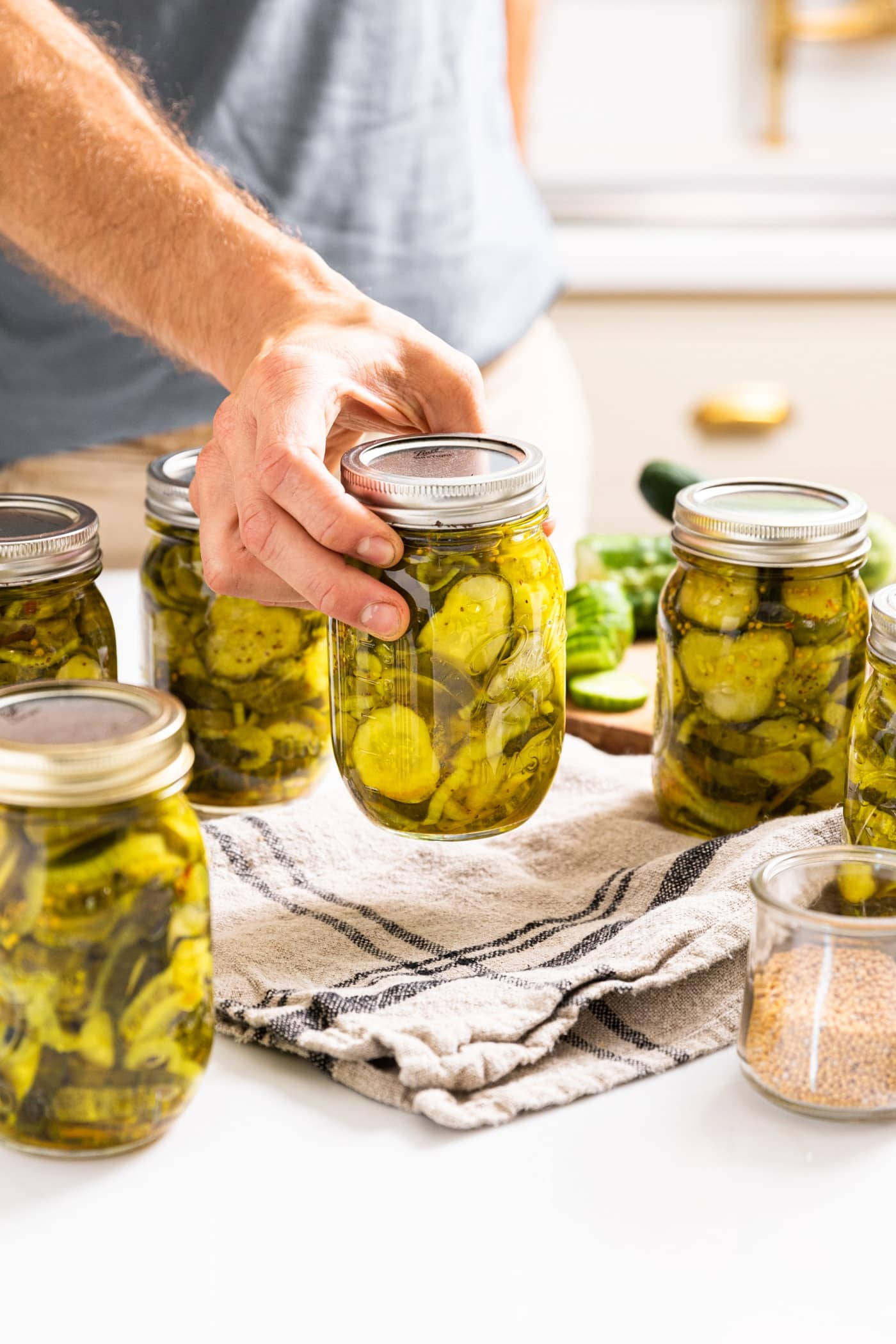 Pickle 桌布 壁紙 — 一個人的手將一罐自製蒔蘿泡菜放在條紋廚房毛巾上，旁邊還有其他裝滿的罐子和新鮮黃瓜。