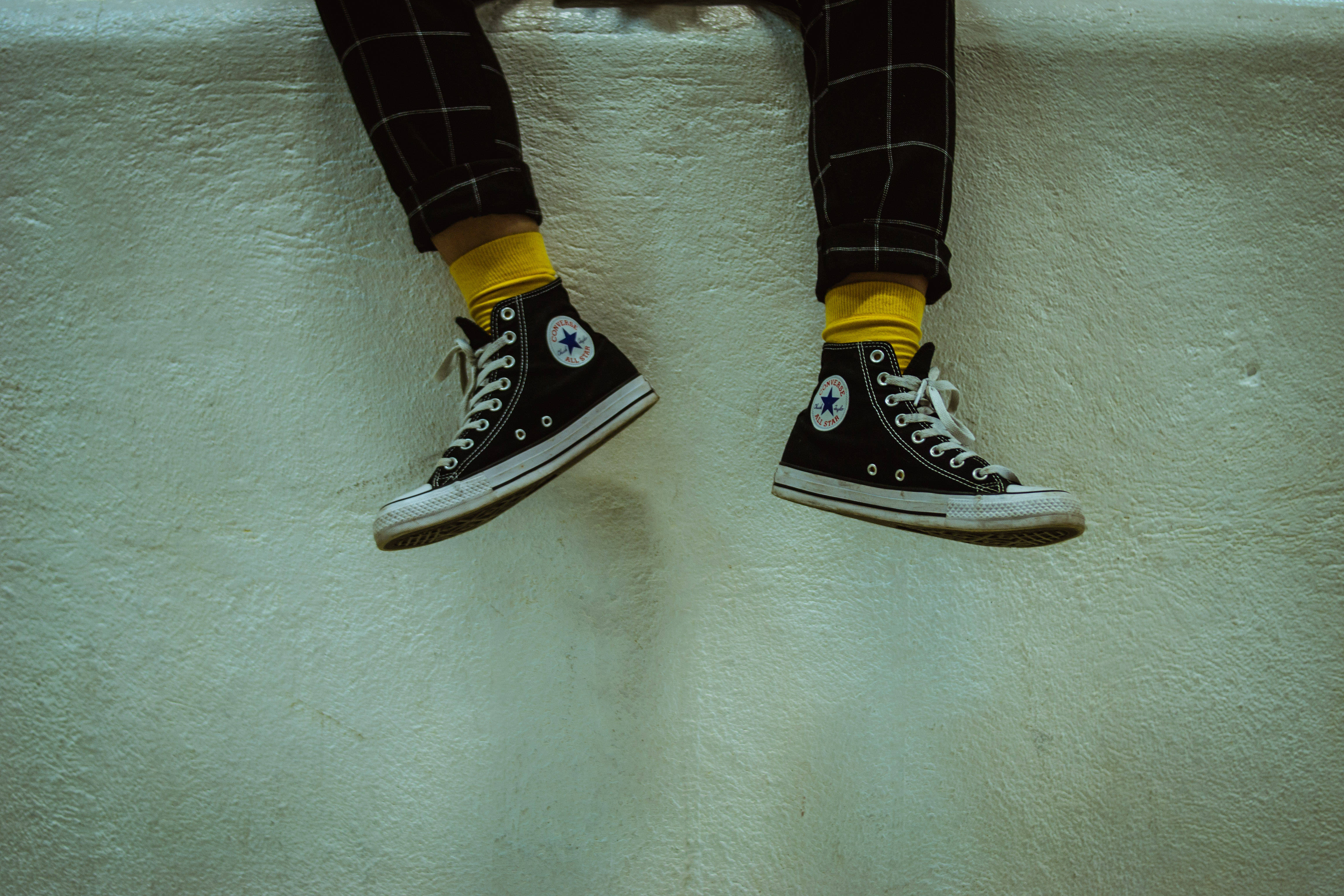 Converse 桌布 壁紙 — 一個人的腳穿著黑色Converse運動鞋和鮮黃色襪子，放鬆地掛在帶有紋理的米白色牆上。