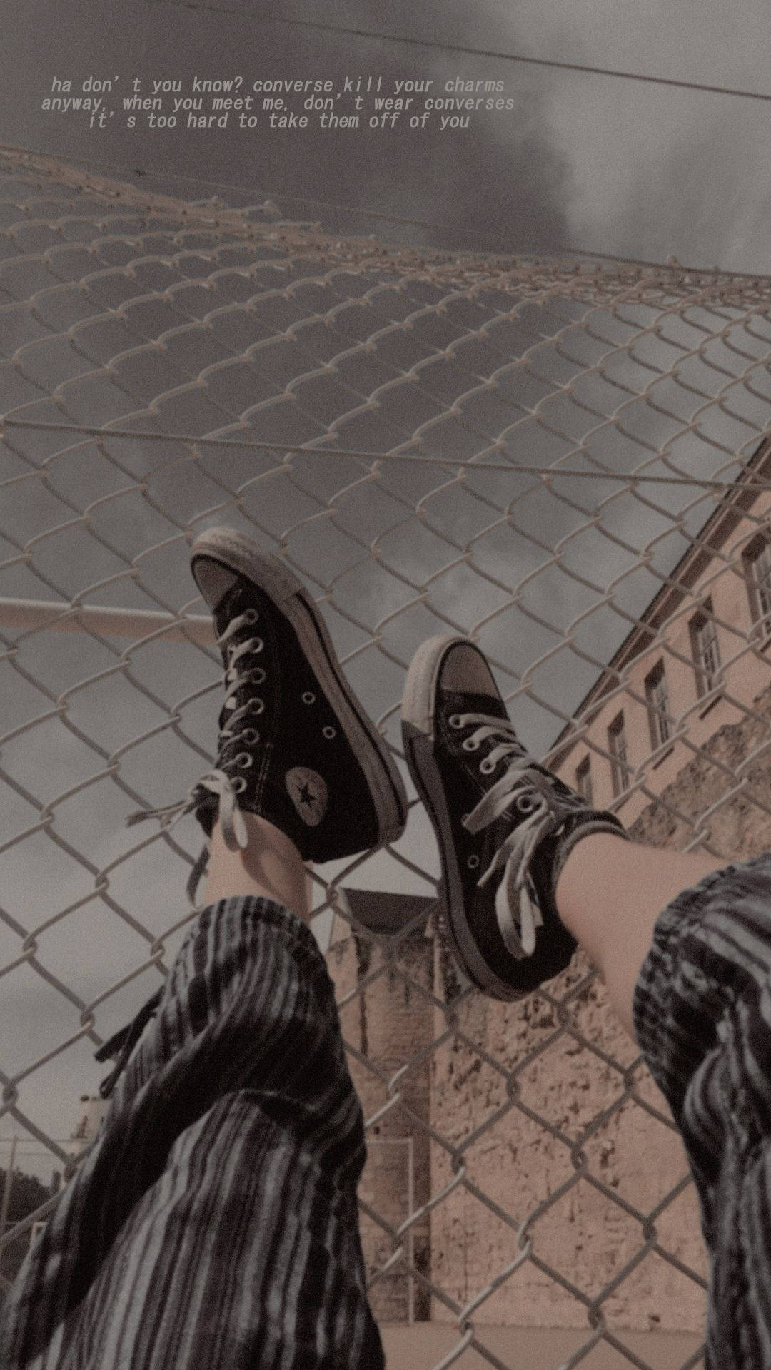 Converse 桌布 壁紙 — 一個人的腳穿著黑色Converse運動鞋，靠著鐵絲網圍欄，背景是粗糙的城市景觀，並覆蓋著詩意的文字。