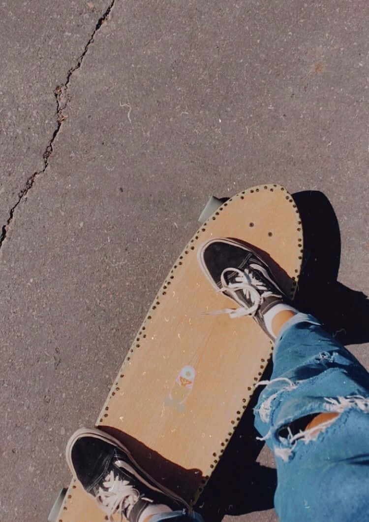 Retro Skateboard 桌布 壁紙 — 一個人的腳穿著黑色運動鞋和破洞牛仔褲，站在木製長板滑板上，下方是裂開的瀝青街道。