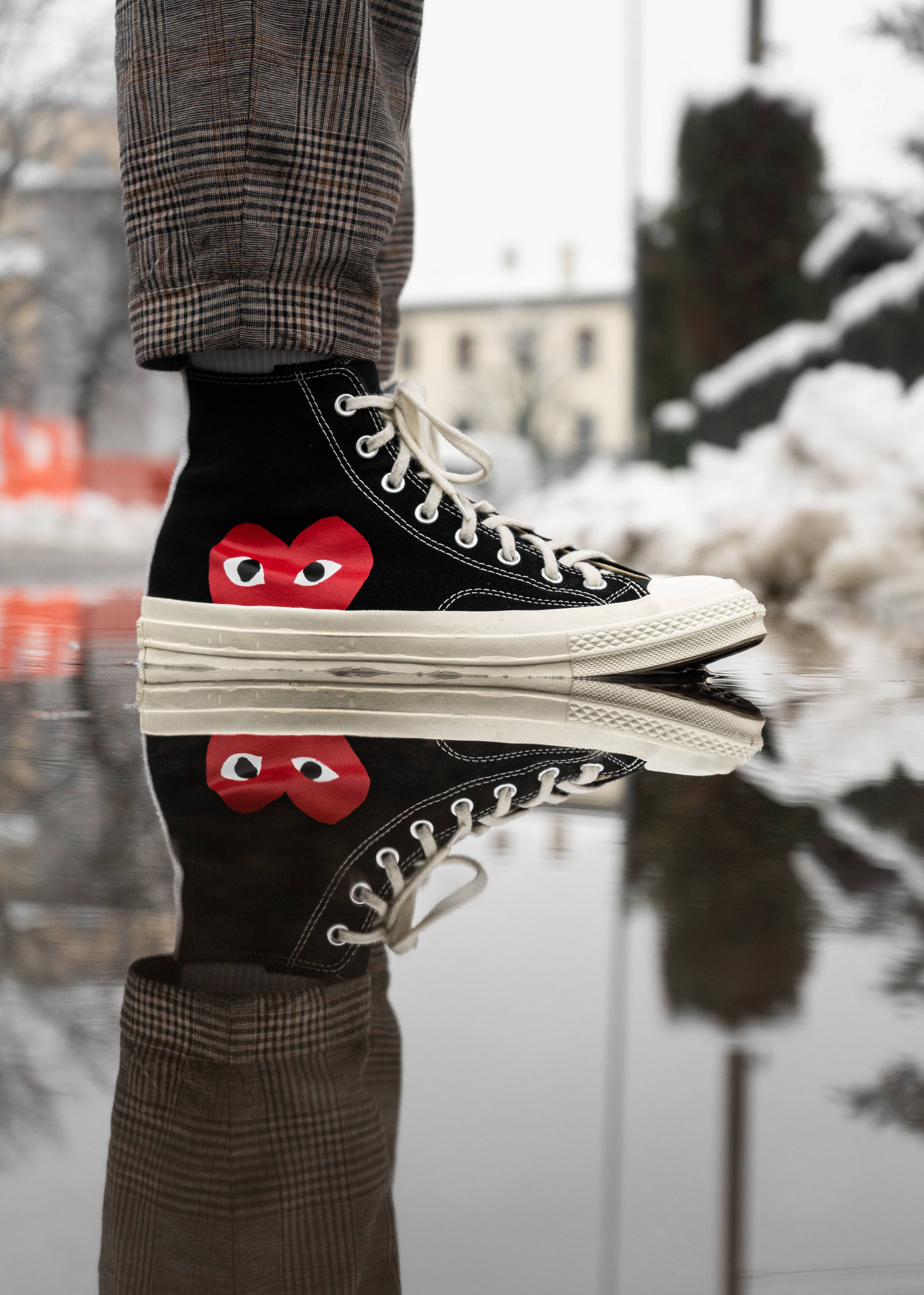 Converse 桌布 壁紙 — 一個人的腳穿著黑色高筒運動鞋，上面有紅色心形和眼睛圖案，站在一個倒映著雪景城市場景的水坑中。