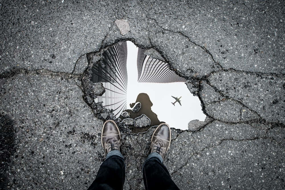 reflection 桌布 壁紙 — 一個人的腳站在裂開的瀝青洞邊緣，露出超現實的顛倒城市景觀，有摩天大樓和飛行的飛機。