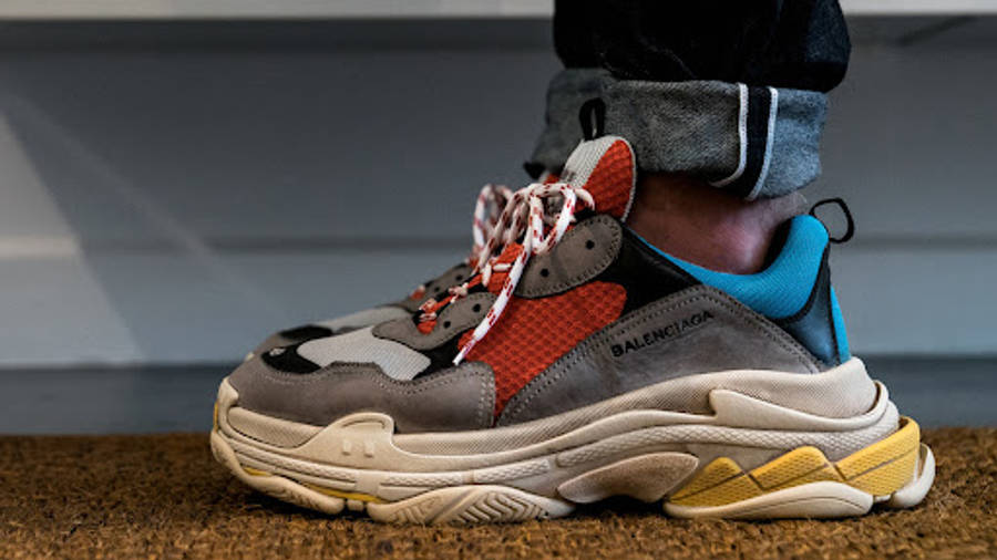 Balenciaga 桌布 壁紙 — 一個人的腳部特寫，穿著色彩繽紛的Balenciaga Triple S運動鞋，站在有紋理的棕色地板上。