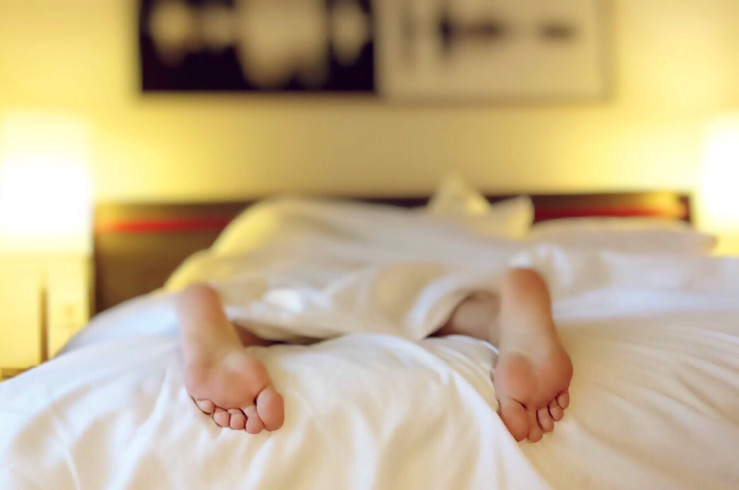 Bedroom 桌布 壁紙 — 一個人的赤腳從床上白色床單下露出來，在一個溫暖燈光、舒適的臥室裡。