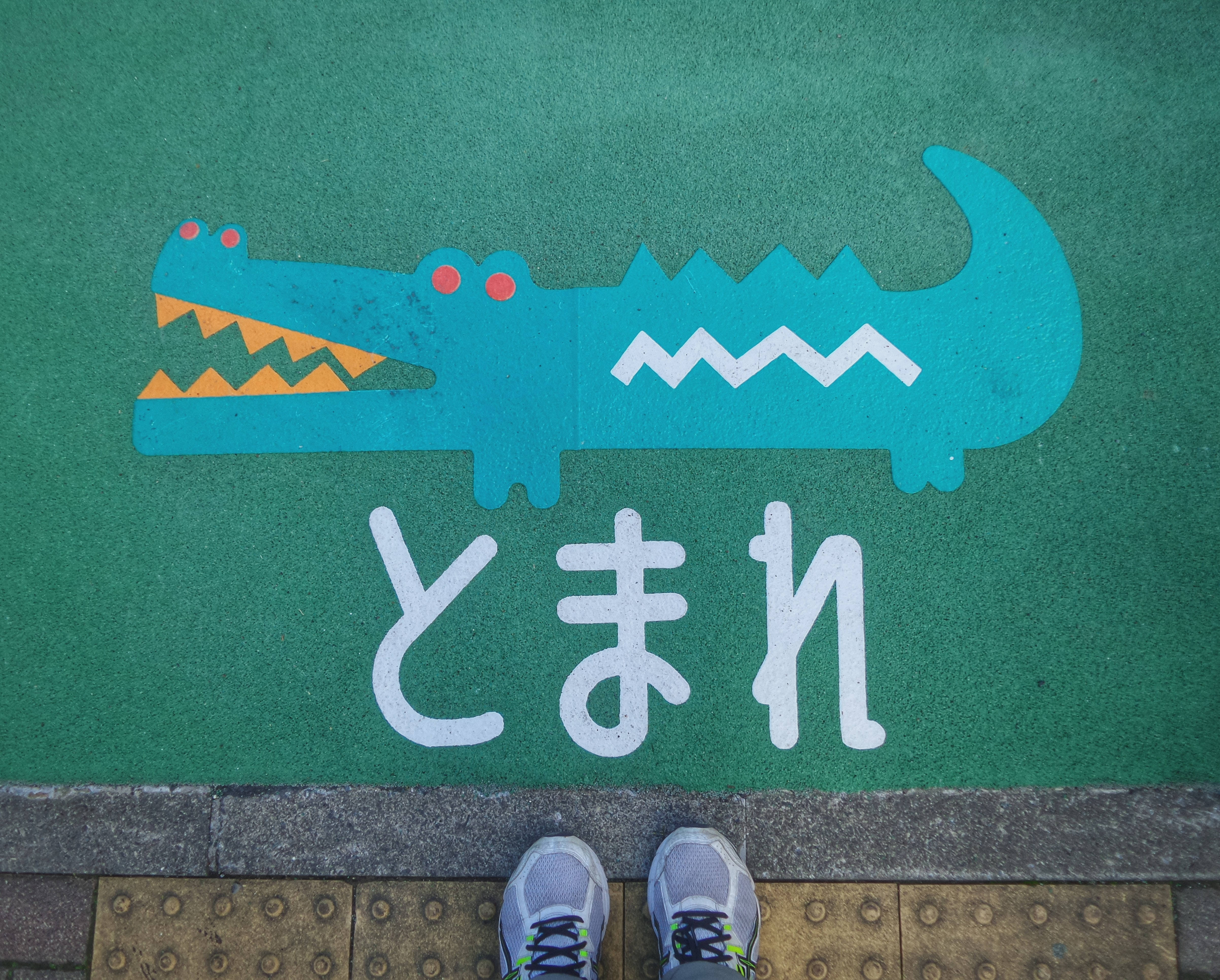 Crocs 桌布 壁紙 — 一個人的運動鞋站在綠色遊樂場表面邊緣，上面有卡通鱷魚和日文字。