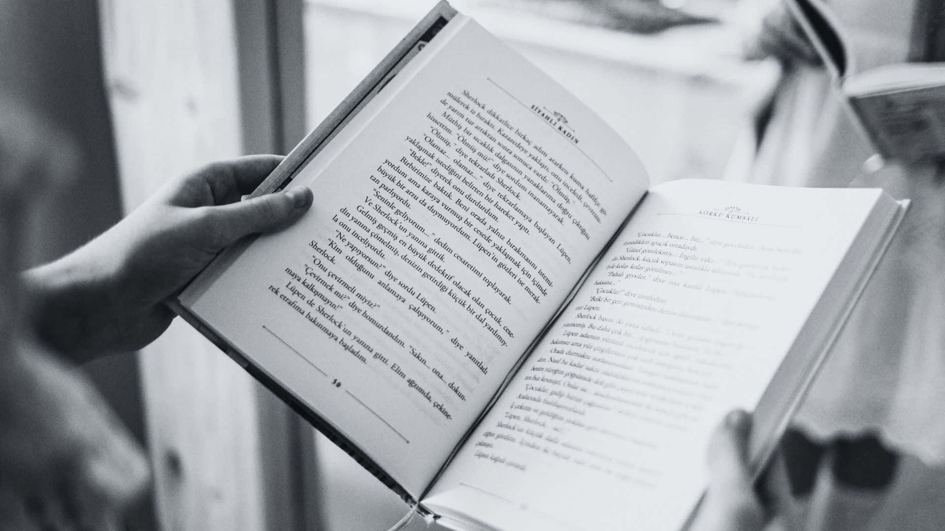Black And White Book 桌布 壁紙 — 一個人的雙手捧著一本打開的書，書頁上文字密集，背景柔和模糊。
