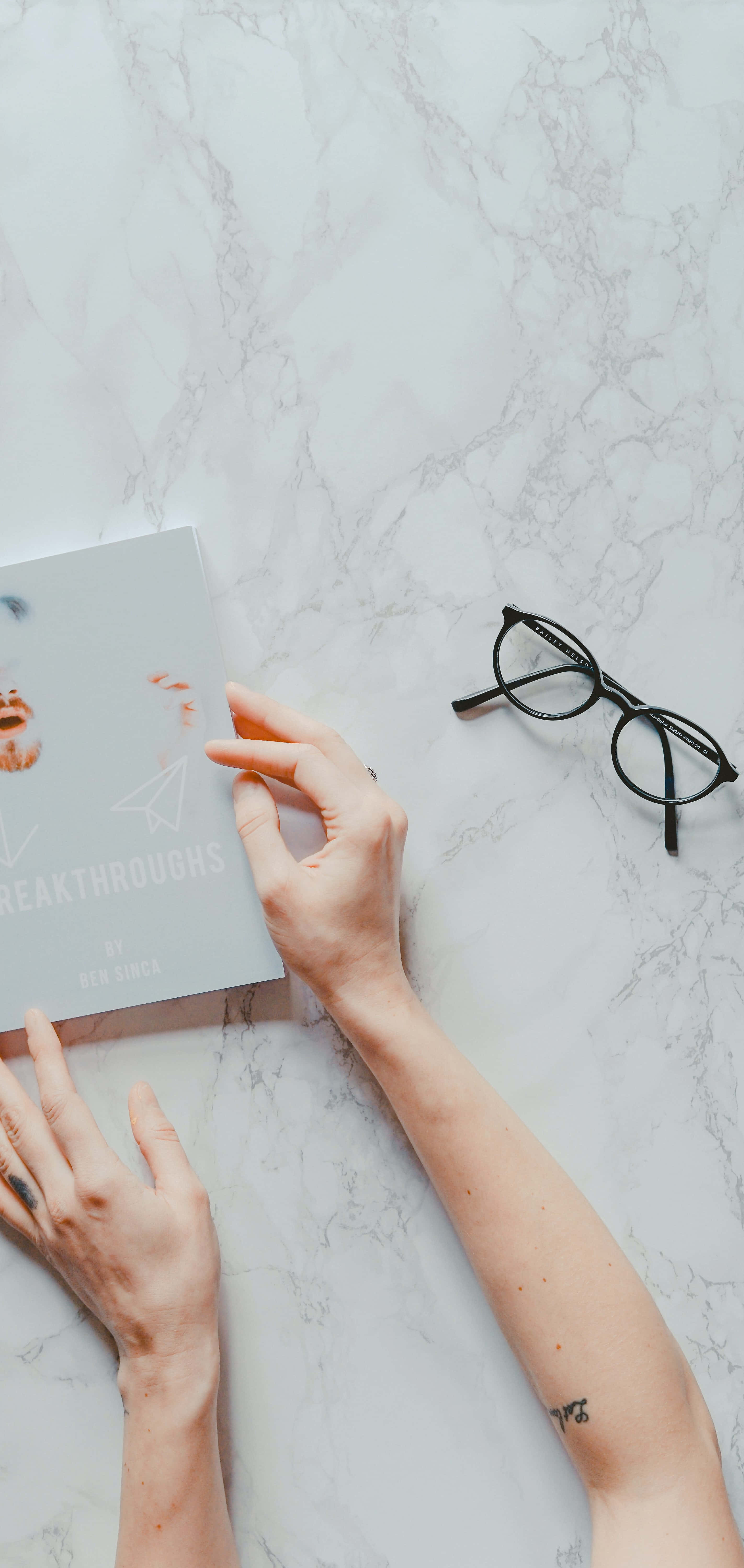 抽象 桌布 壁紙 — 一個人的雙手放在一本名為「Breakthroughs」的書上，旁邊有一副黑框眼鏡，置於白色大理石表面。