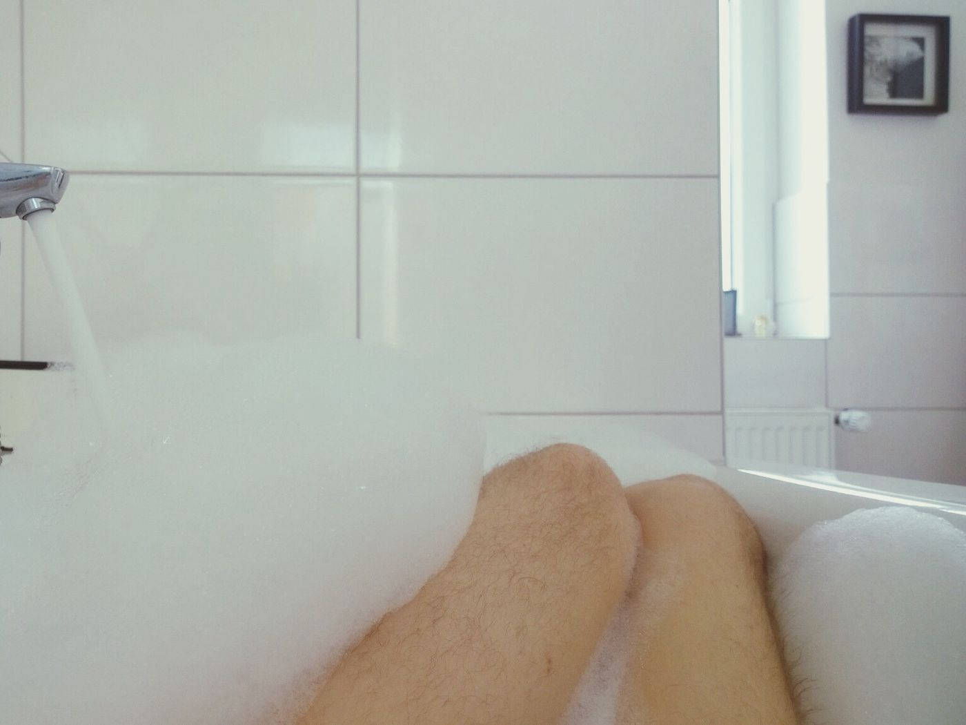 Bathtub 桌布 壁紙 — 一個人的雙腿在充滿泡泡的浴缸中放鬆，水從鉻合金水龍頭流出，濺在白色瓷磚牆上。