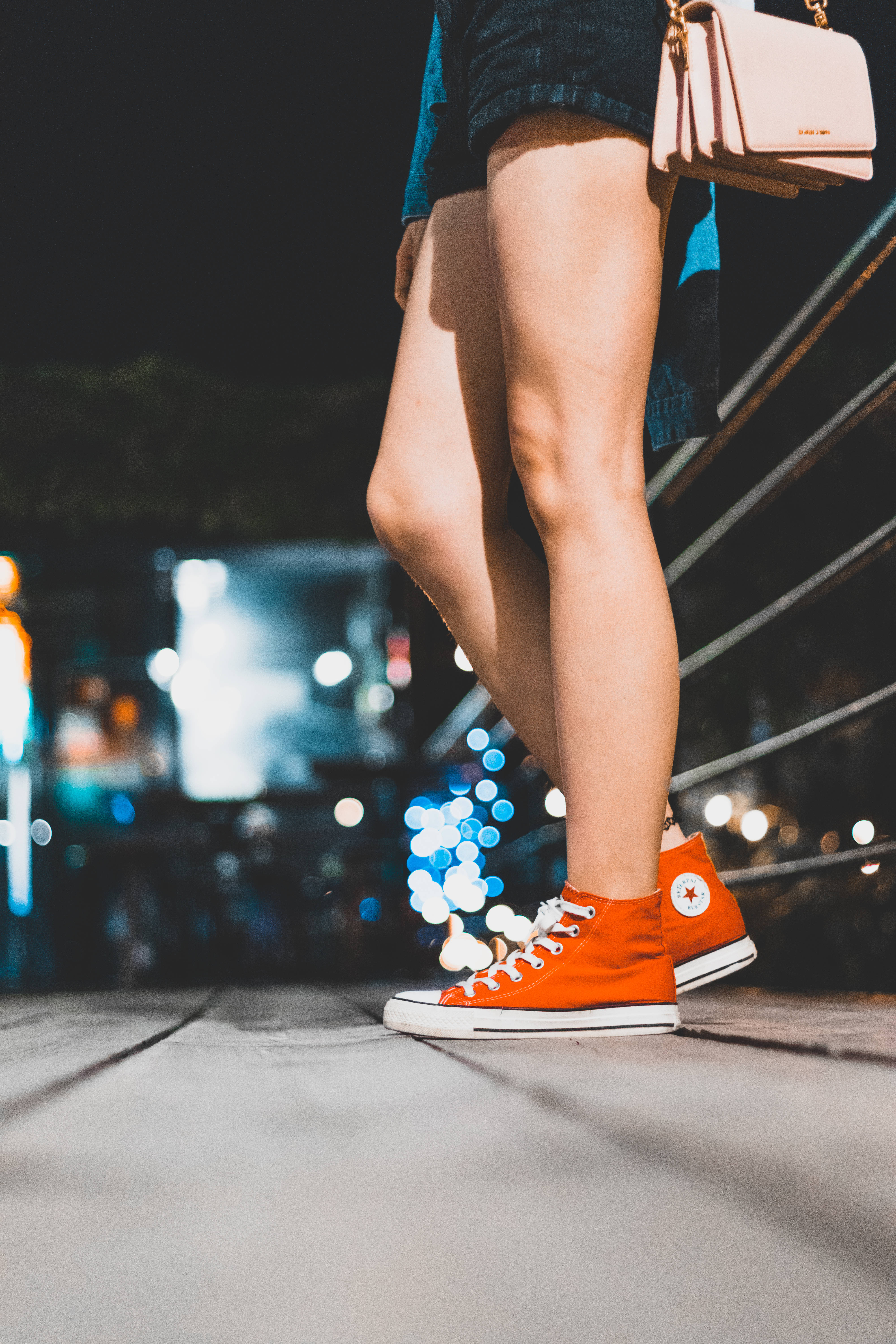 Converse 桌布 壁紙 — 一個人的雙腿穿著亮橙色運動鞋，夜晚站在木製步道上，背後是模糊的城市燈光。
