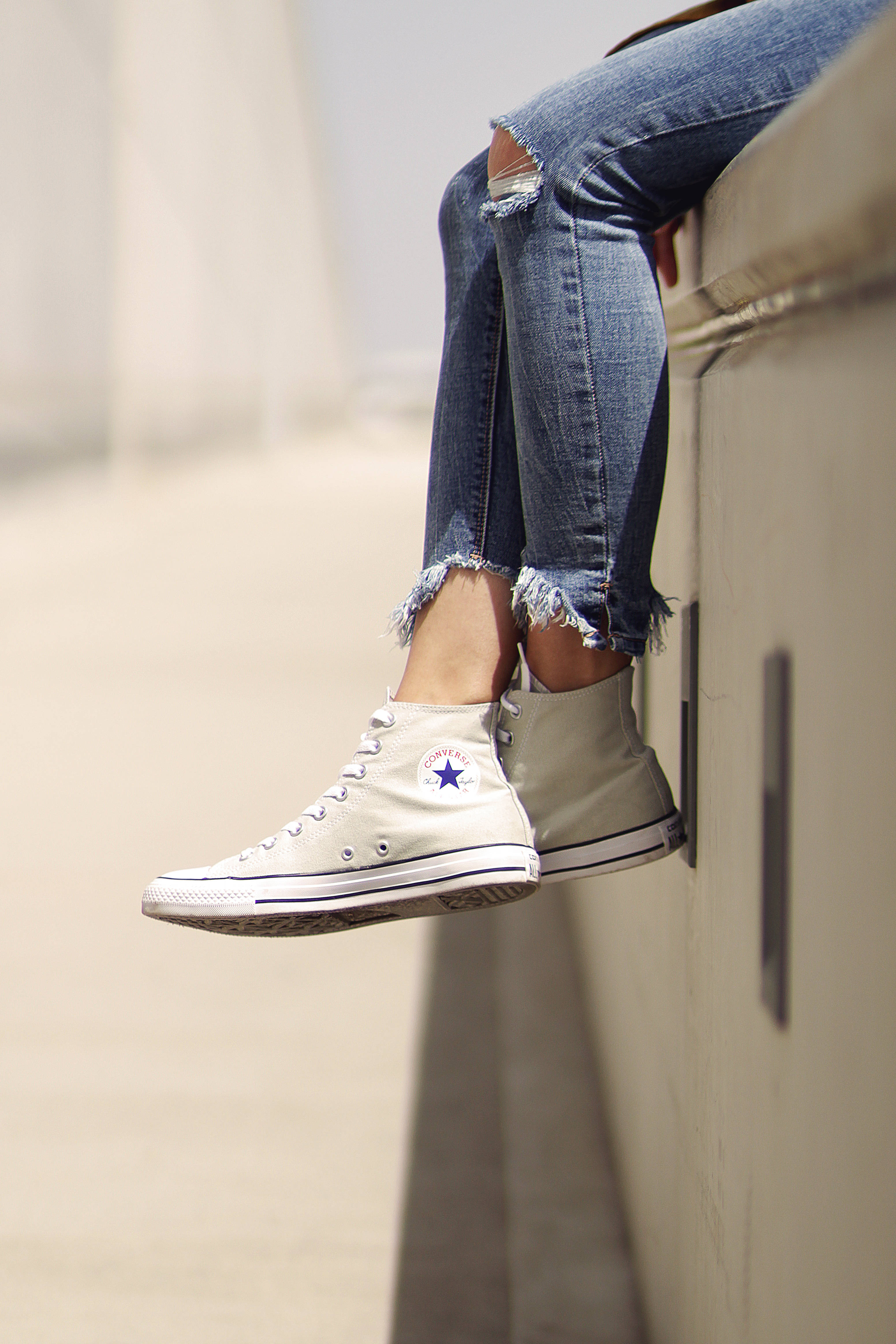 Converse 桌布 壁紙 — 一個人的雙腿穿著破洞牛仔褲和白色高筒運動鞋，在明亮的日光下隨意地懸掛在混凝土邊緣上。