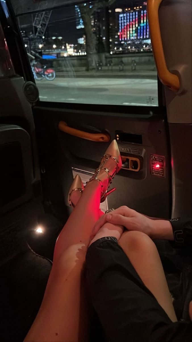 情侶 桌布 壁紙 — 一個人的雙腿穿著金色細高跟鞋，擱在汽車座位上，一隻手輕輕觸碰大腿，夜間窗外閃爍著充滿活力的城市景觀。