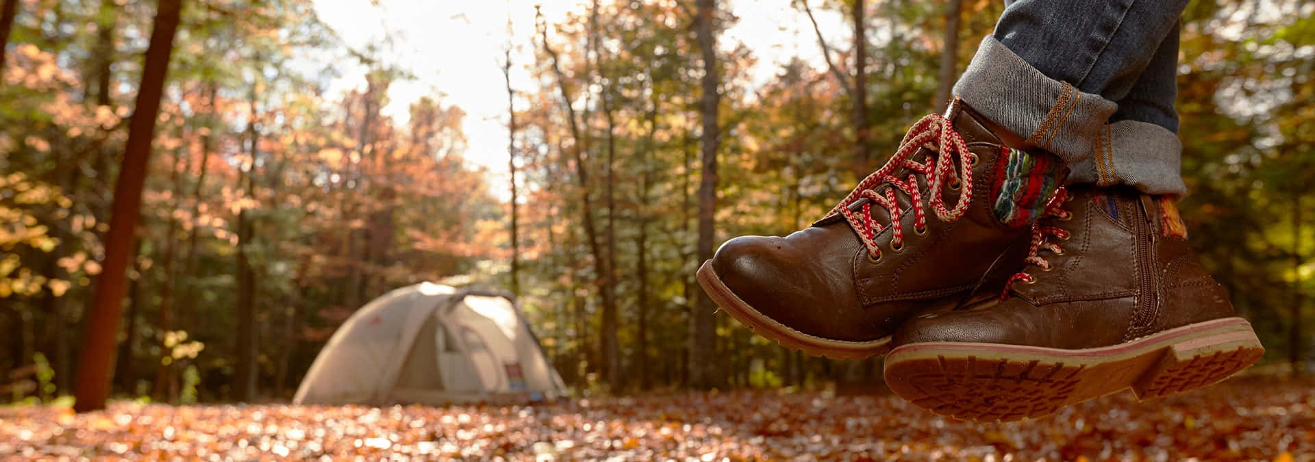 Fall Camping 桌布 壁紙 — 一個人的靴子懸掛在鋪滿秋葉的森林地面上方，背景中有一個帳篷。