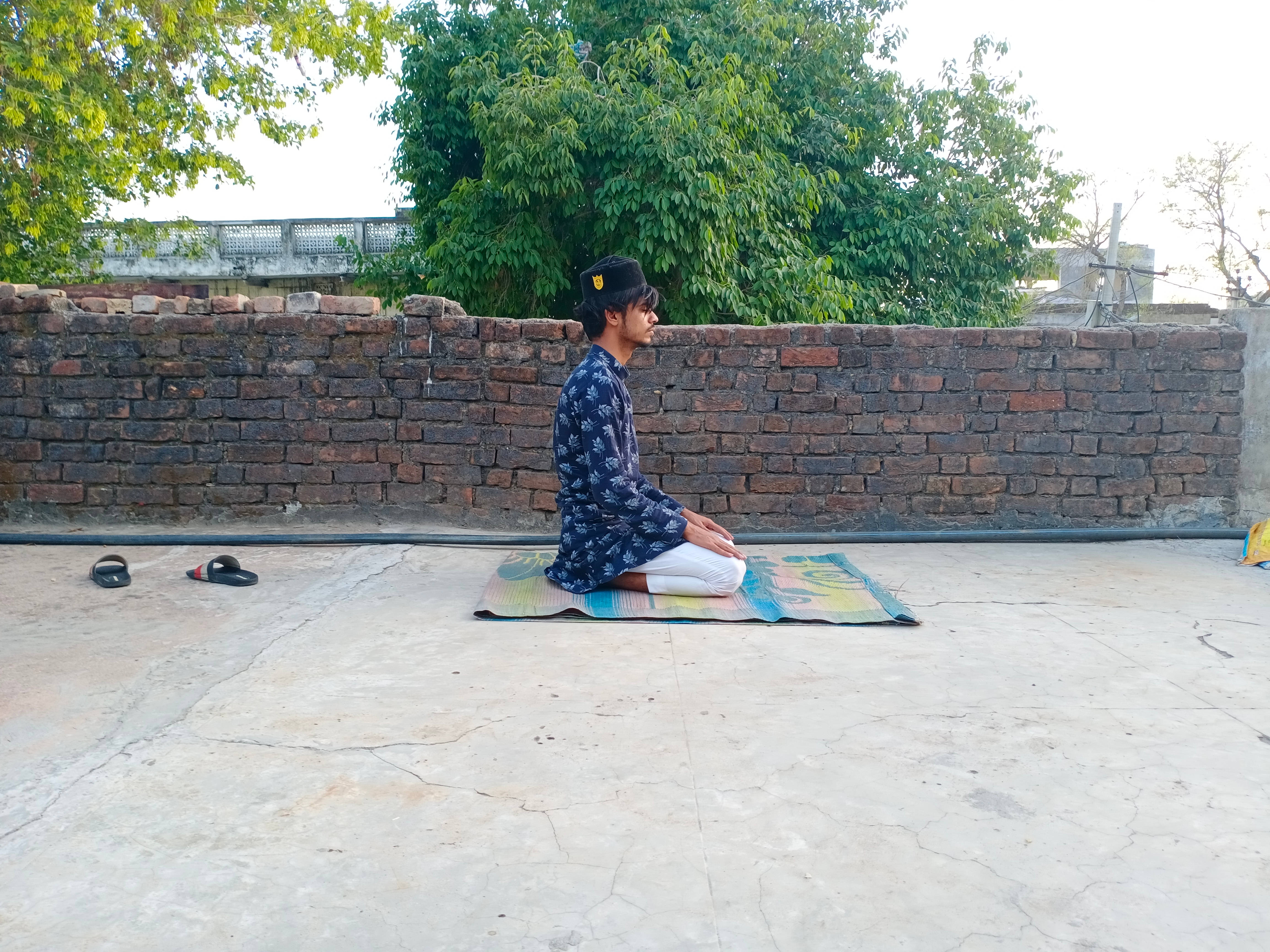 Ajmer 桌布 壁紙 — 一個人盤腿坐在彩色墊子上，在混凝土屋頂上冥想，周圍有綠樹和磚牆。