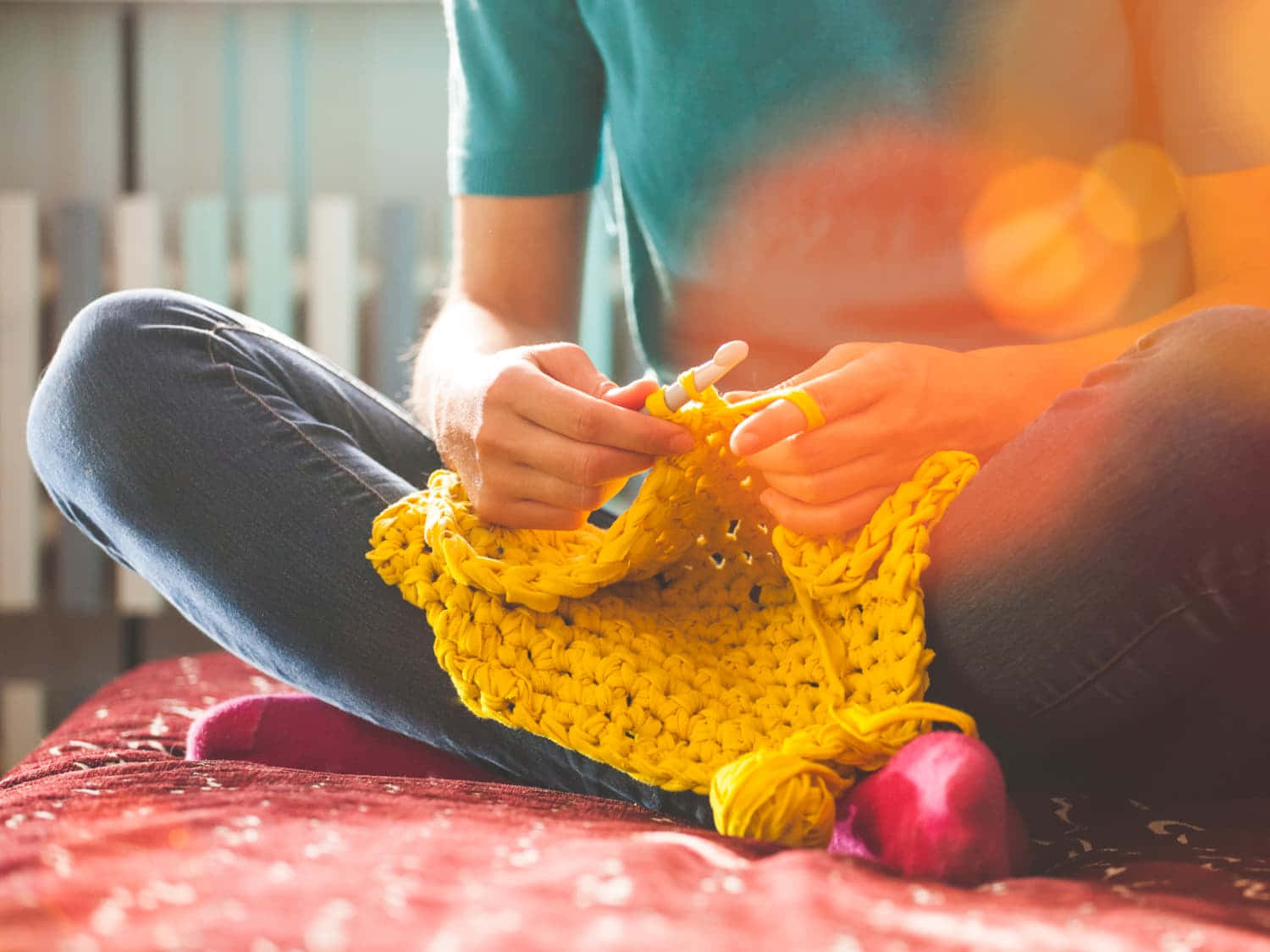 Crocheting 桌布 壁紙 — 一個人盤腿坐在紅色床上，編織一條鮮黃色厚圍巾，溫暖的陽光從他們身後的窗戶照射進來。