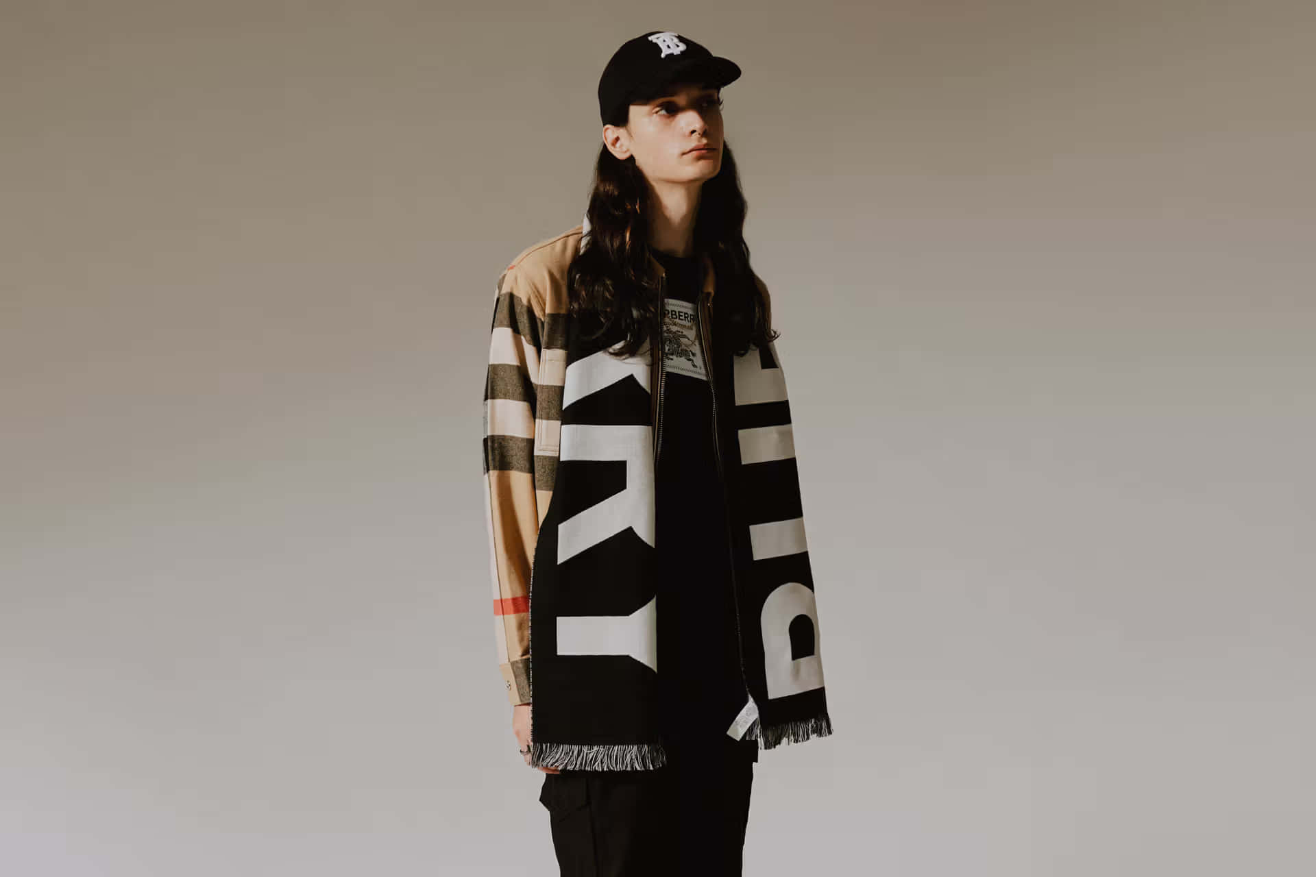 潮流玩家 桌布 壁紙 — 一個人穿著 Burberry 格紋外套和一條帶有粗體白色字樣的大圍巾，站在中性工作室背景前。