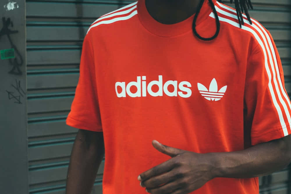 愛迪達 桌布 壁紙 — 一個人穿著一件大膽的橙色adidas T恤，帶有白色條紋和標誌，背景是塗鴉噴漆的金屬百葉窗。