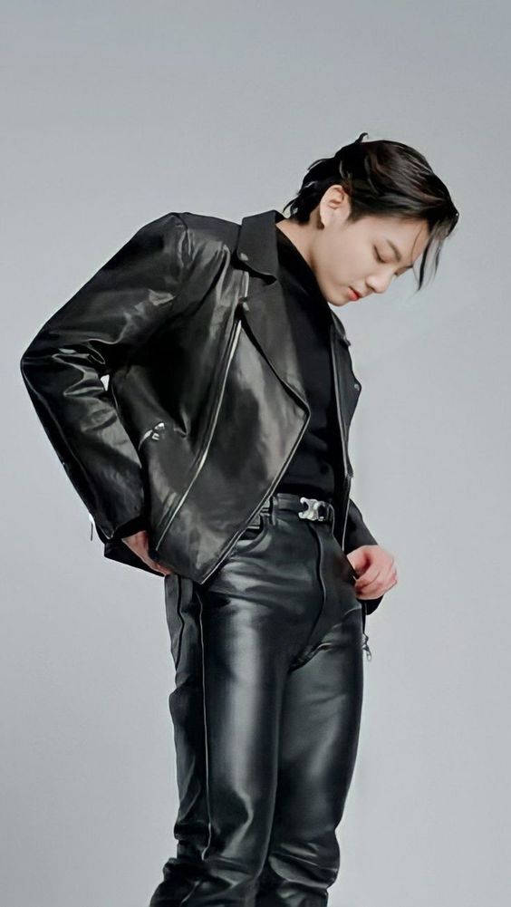 Jungkook 桌布 壁紙 — 一個人穿著亮面黑色皮夾克和皮褲，雙手插袋，在純灰色攝影棚背景前擺姿勢。