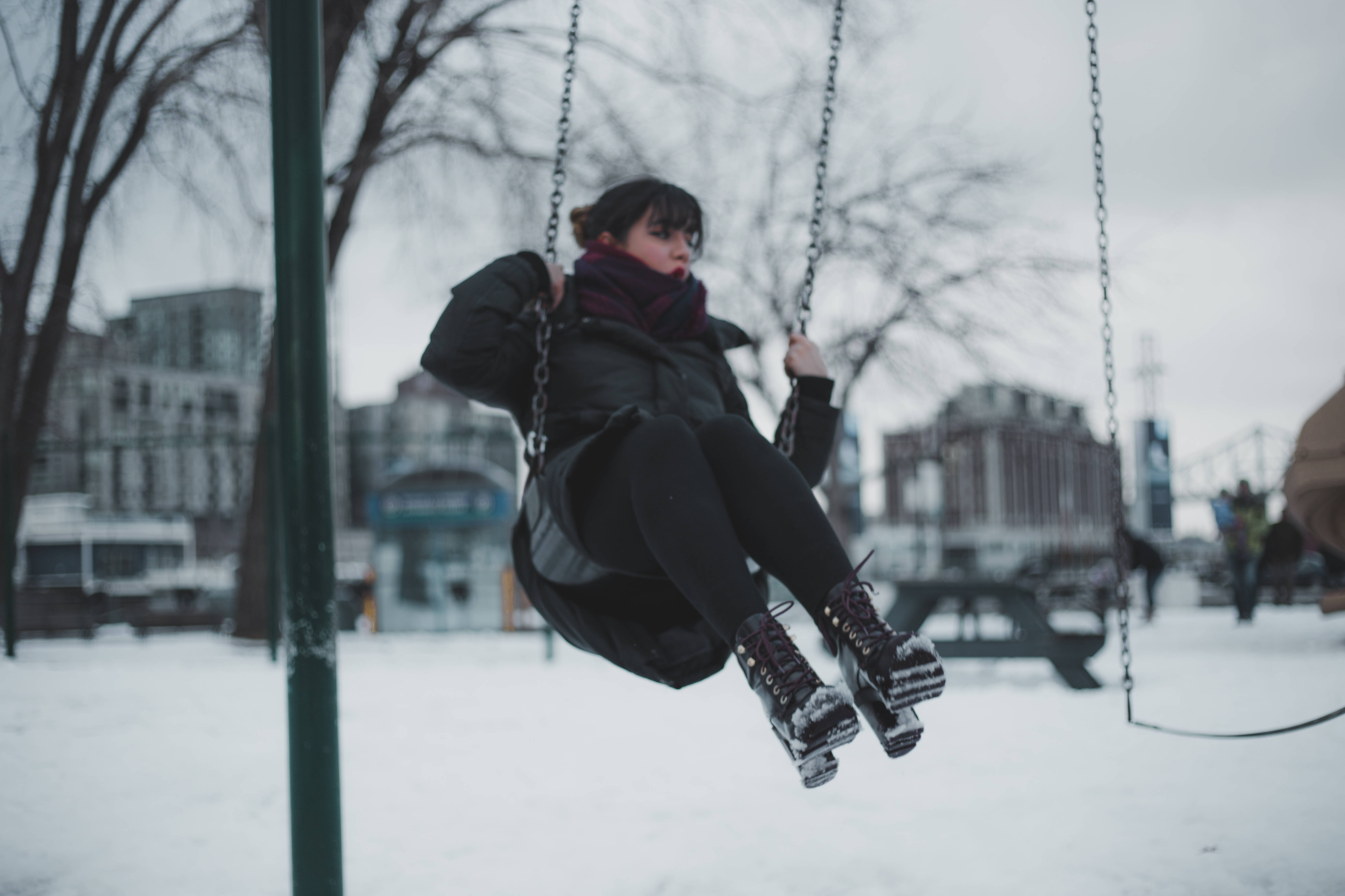 Playground 桌布 壁紙 — 一個人穿著冬季裝備坐在鞦韆上，背景是白雪覆蓋的城市公園，有光禿禿的樹木和建築物。