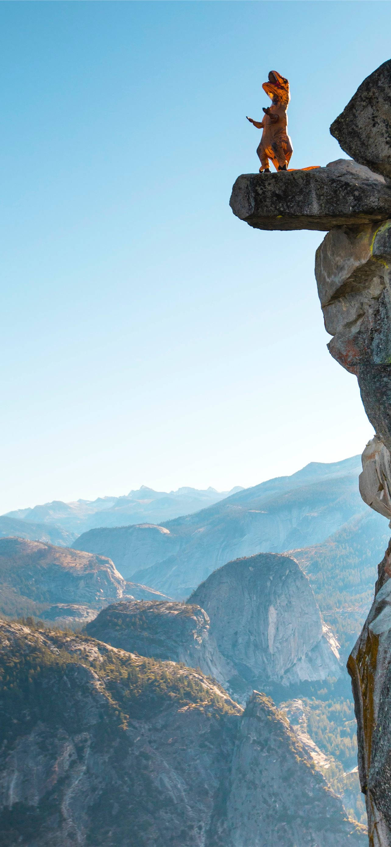 Climbing 桌布 壁紙 — 一個人穿著恐龍服裝站在岩石懸崖上，俯瞰著廣闊的山谷，天空清澈湛藍。