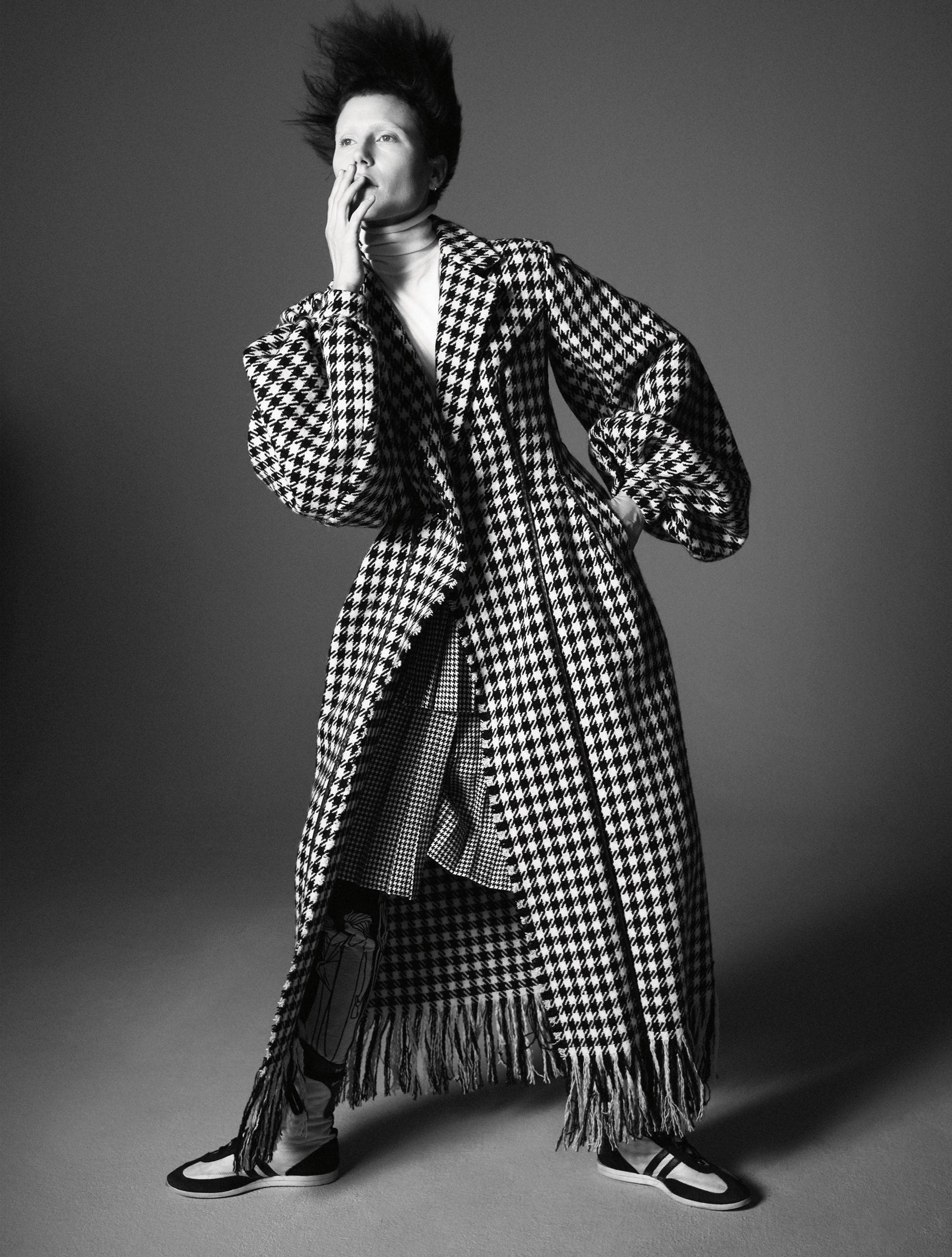 Balenciaga 桌布 壁紙 — 一個人穿著戲劇性的千鳥格大衣，帶有流蘇下擺和超大袖子，在純灰色工作室背景前擺姿勢。