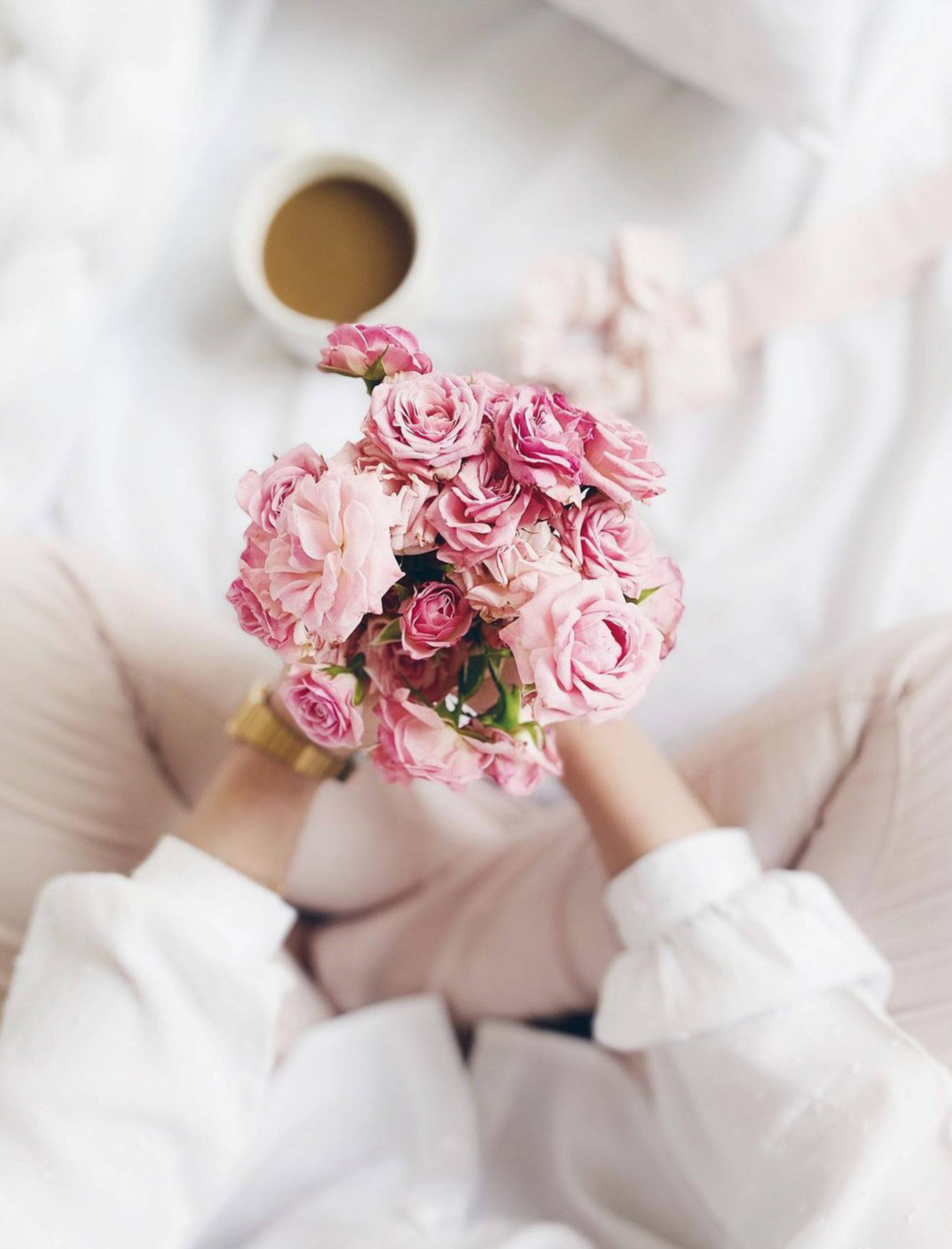 Aesthetic Rose 桌布 壁紙 — 一個人穿著柔軟的白色衣服，坐在床上，手裡拿著一束粉紅玫瑰，旁邊有一杯咖啡。