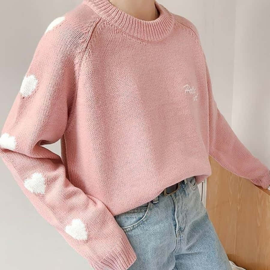 Pink Sweater 桌布 壁紙 — 一個人穿著柔軟的粉紅色針織毛衣，帶有白色心形圖案和淺藍色牛仔褲，背景為純色。