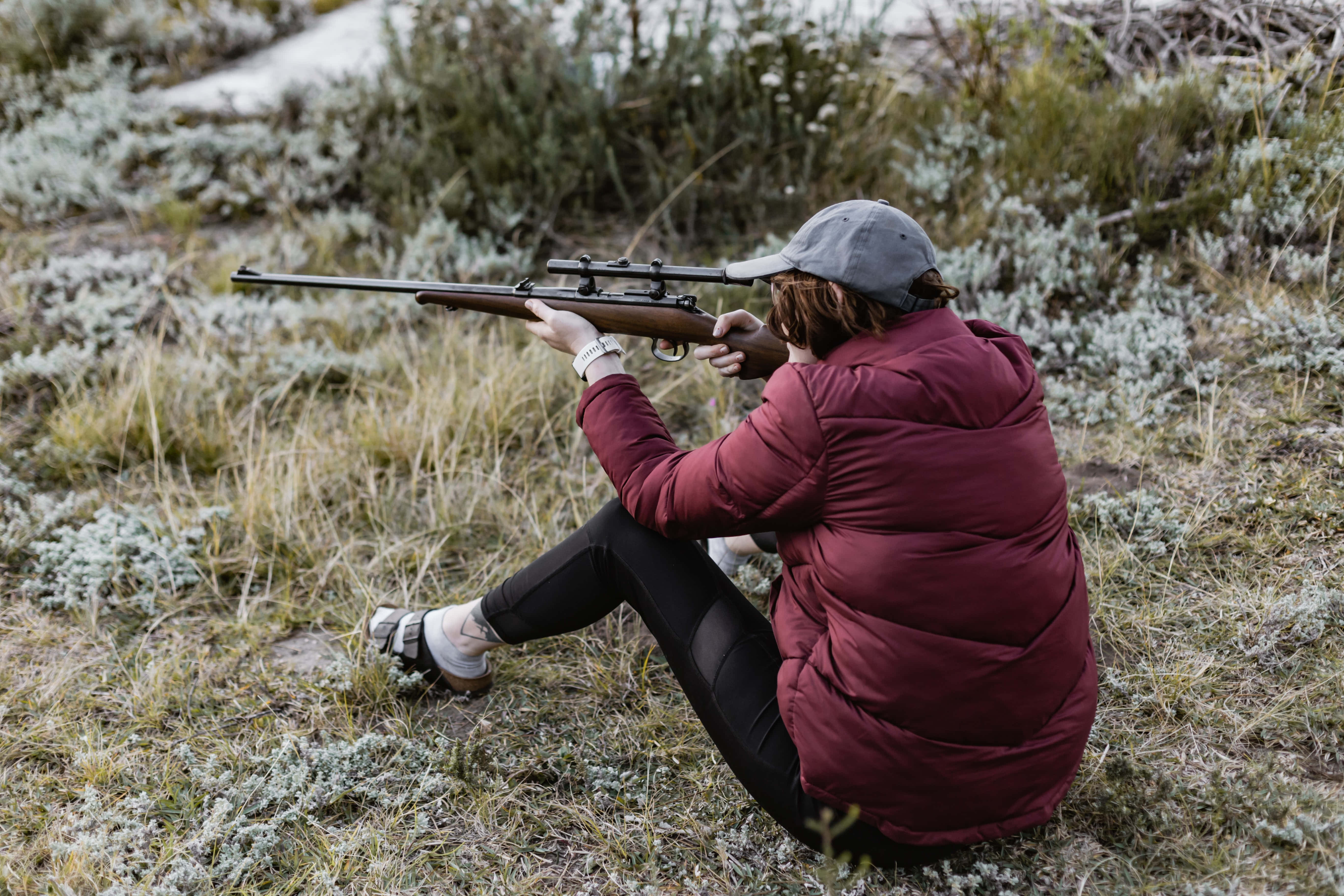 Rifle 桌布 壁紙 — 一個人穿著栗色外套，盤腿坐在草地上，專注地瞄準步槍。