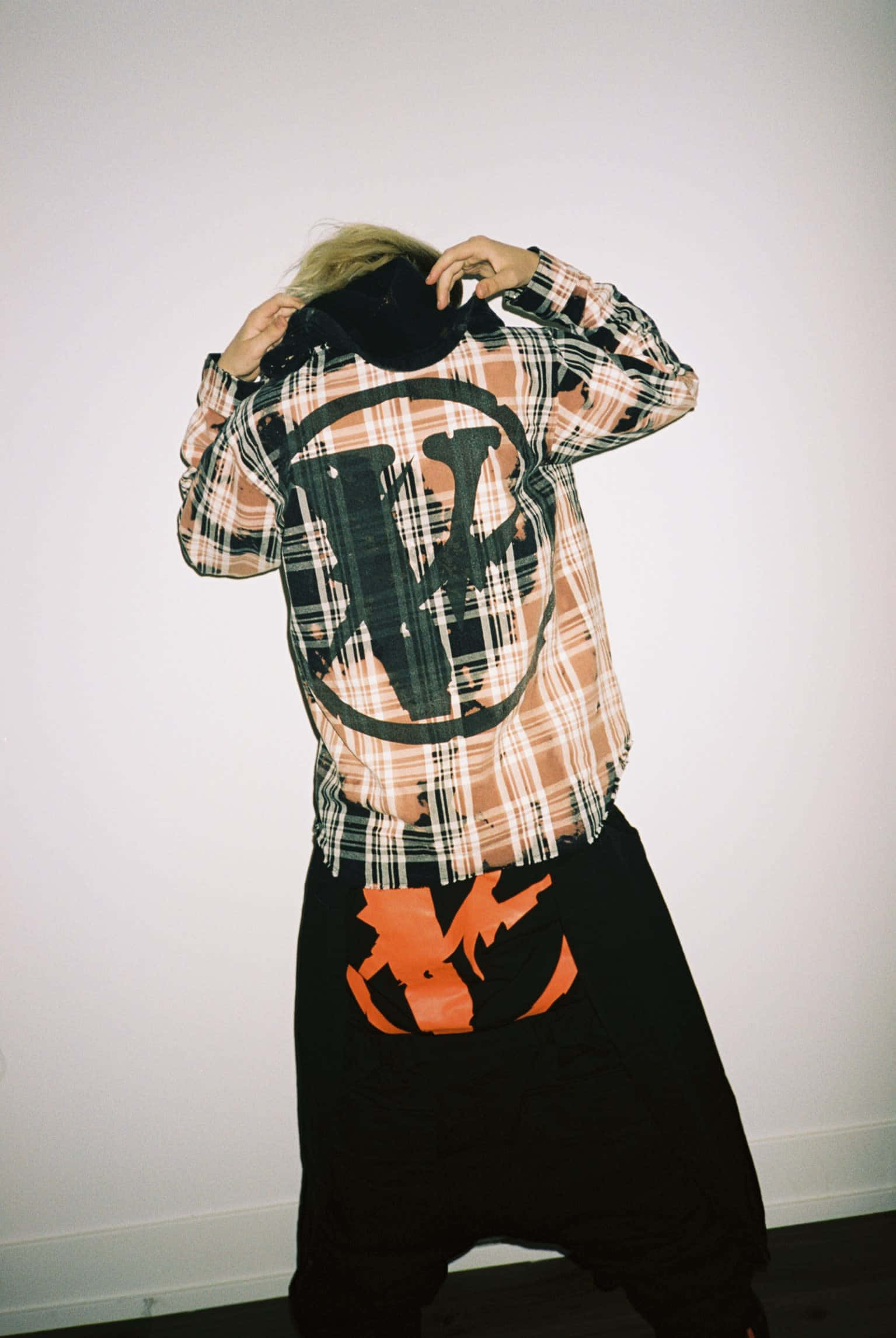 Vlone 桌布 壁紙 — 一個人穿著格紋襯衫，上面有大型黑色標誌和磨邊下擺，轉身背對，雙手調整帽子，背景是純白牆壁。