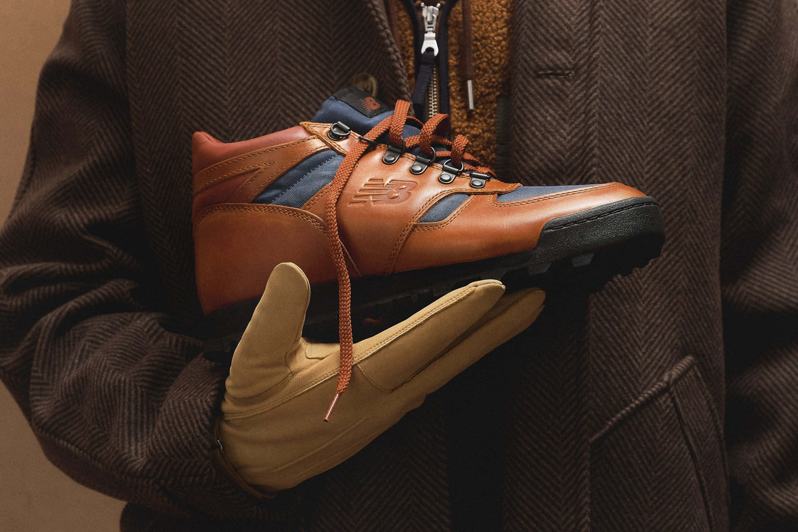 New Balance 桌布 壁紙 — 一個人穿著棕色人字紋外套和棕褐色手套，手持一雙帶橙色鞋帶的棕色和藍色New Balance遠足鞋。