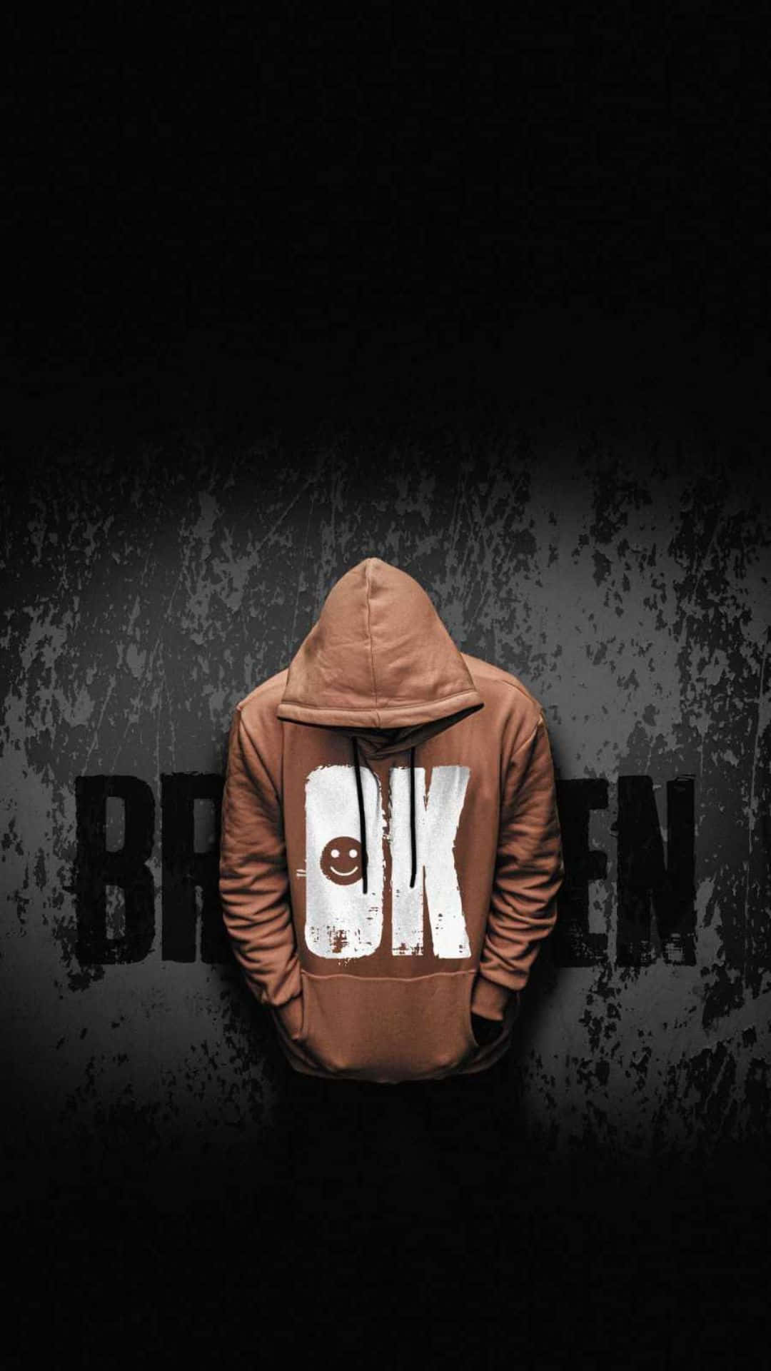 Broken 桌布 壁紙 — 一個人穿著棕色連帽衫，背面有笑臉圖案，背對一面黑暗粗糙的牆壁，牆上 partial 可見「broken」這個字。