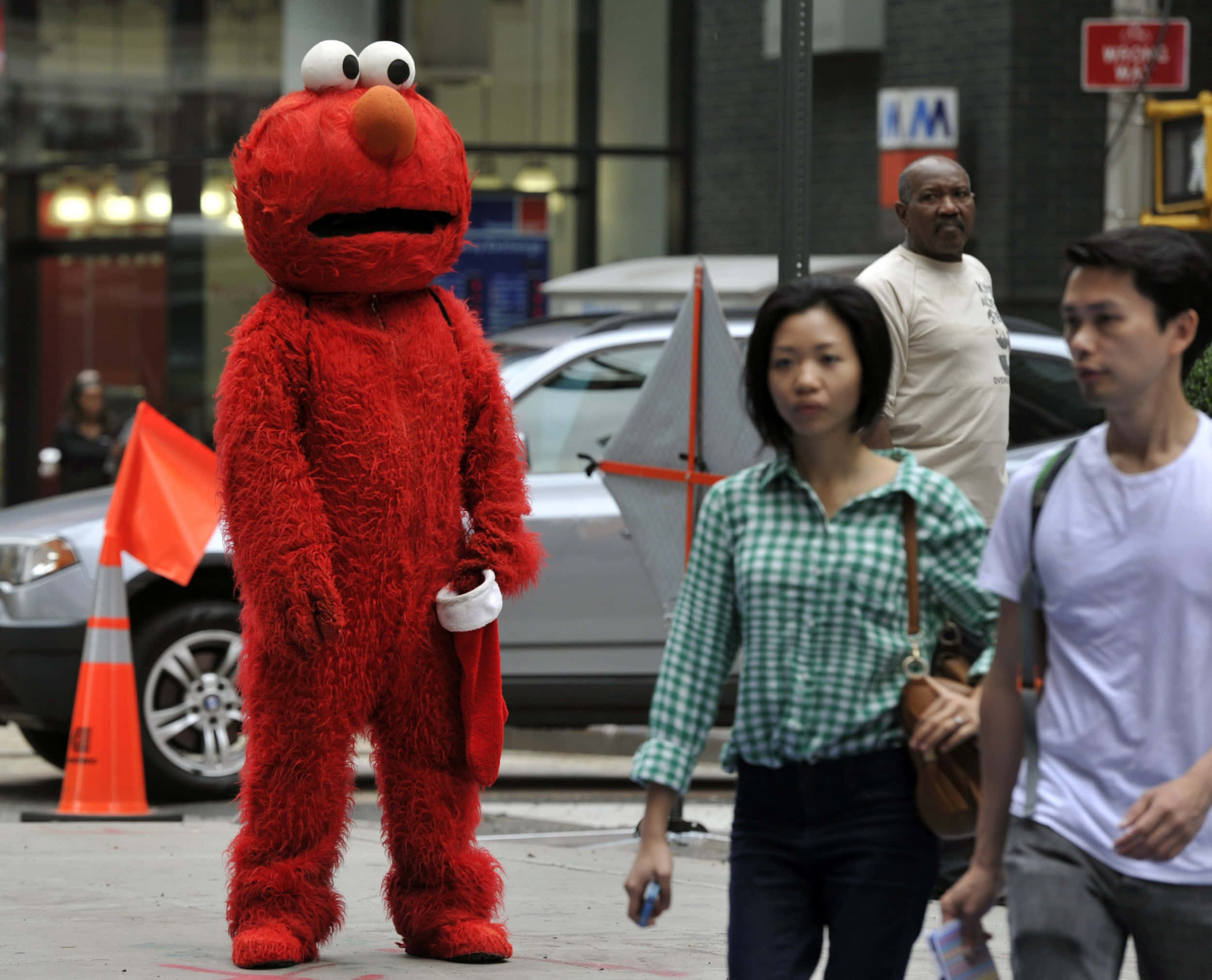 Elmo 桌布 壁紙 — 一個人穿著毛茸茸的紅色Elmo服裝站在城市人行道上，行人走過。